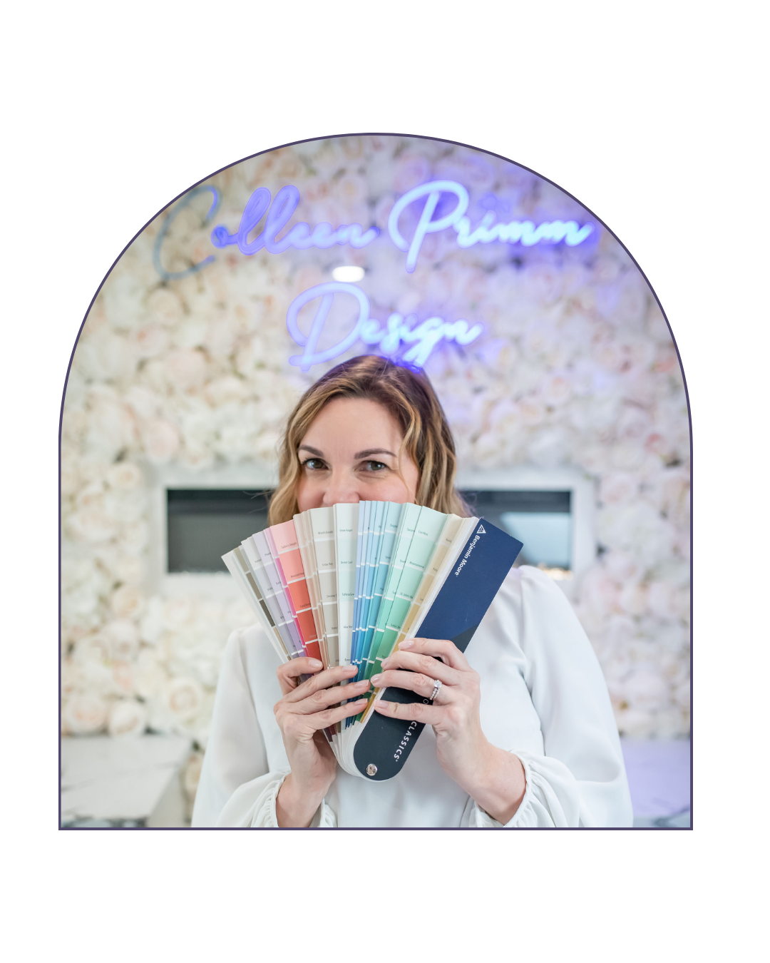 A woman with blonde hair holding fan of paint swatches in front of her face, standing in front of a floral wall with a neon sign that says 'Collen Primm Design'.
