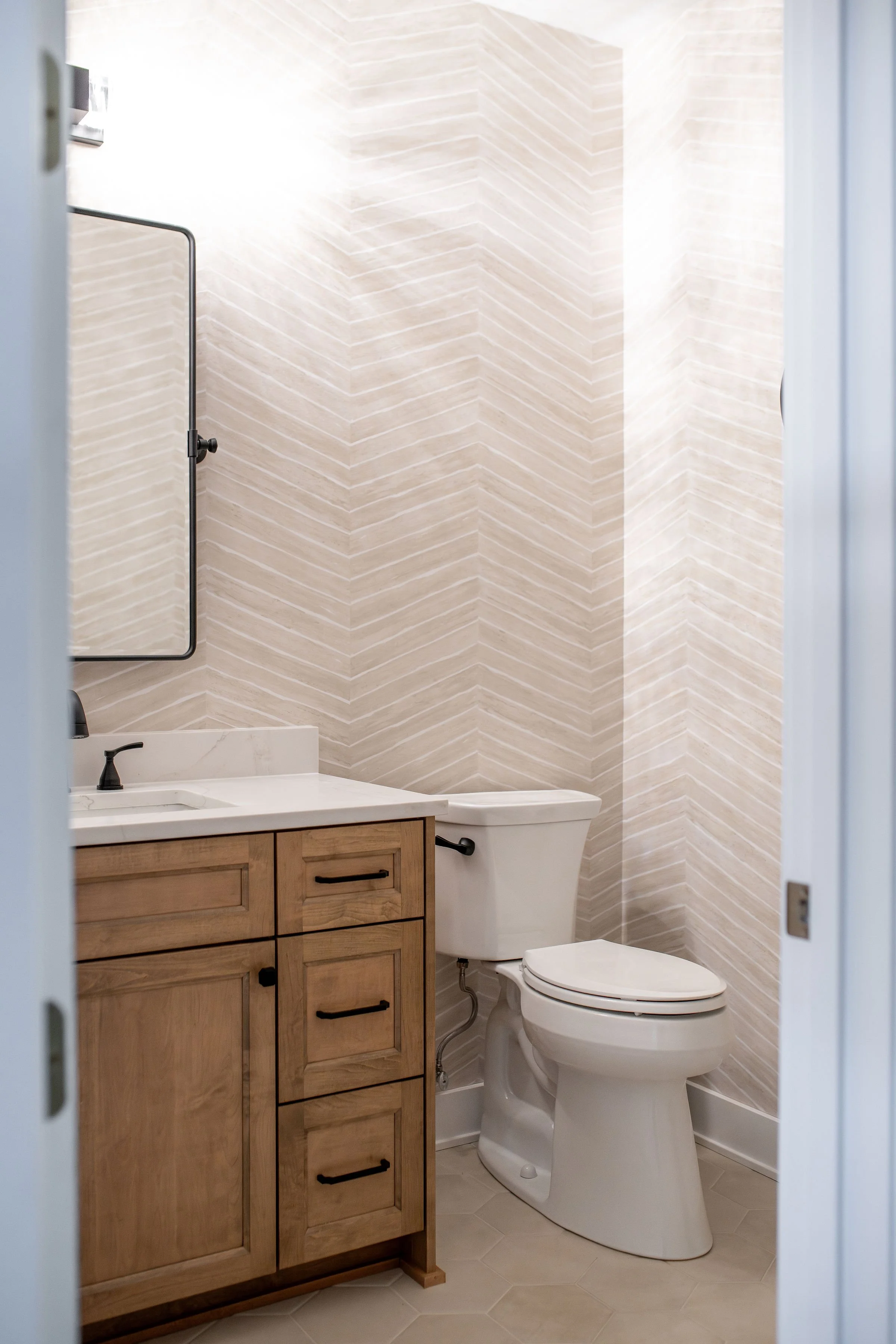 A modern bathroom with a wooden vanity, white countertop, wall-mounted mirror, and a white toilet against a textured beige wall.