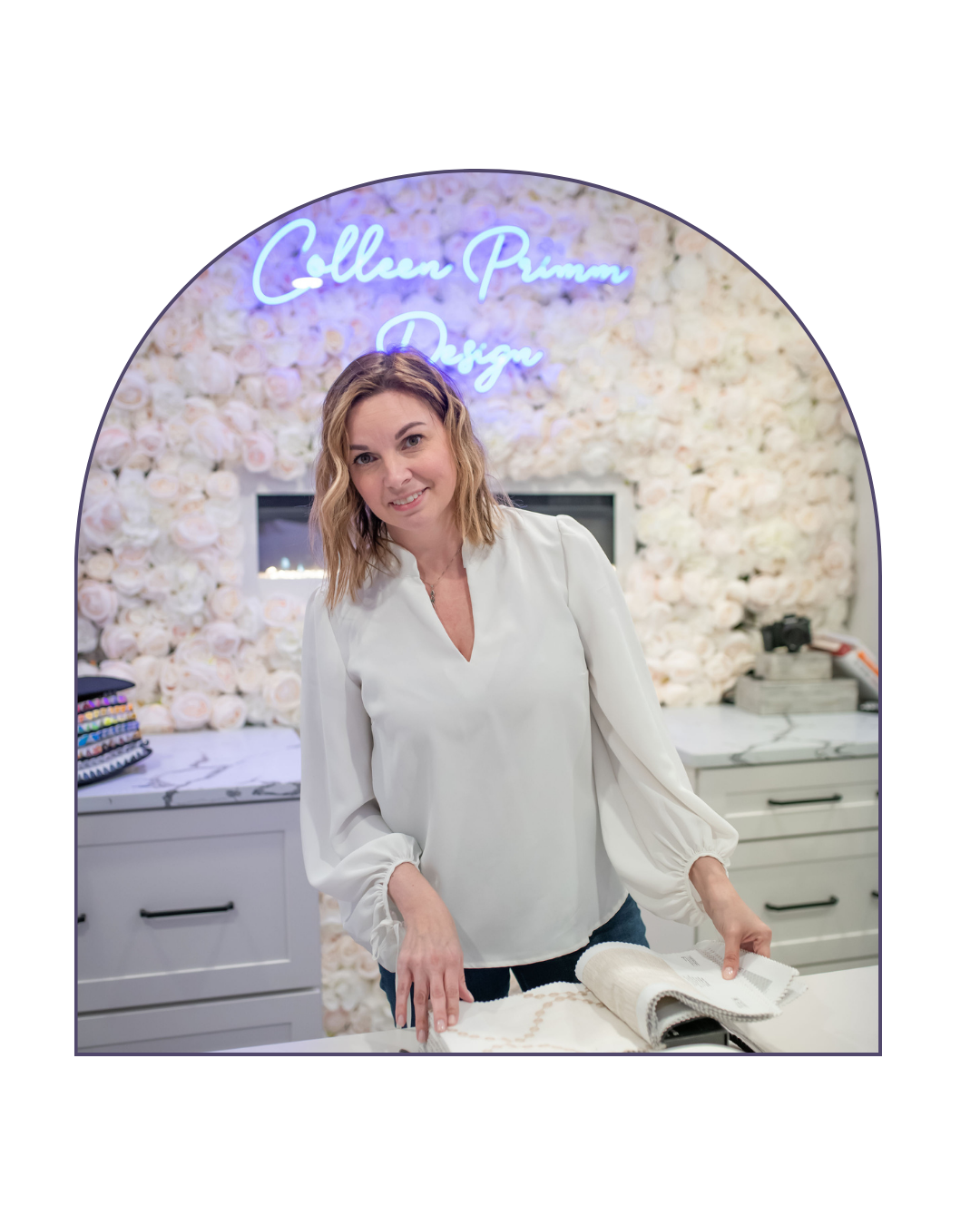 Woman standing at a table with fabric sample book, inside a floral decorated room with a neon sign that reads 'Gollen Prumm Design'.