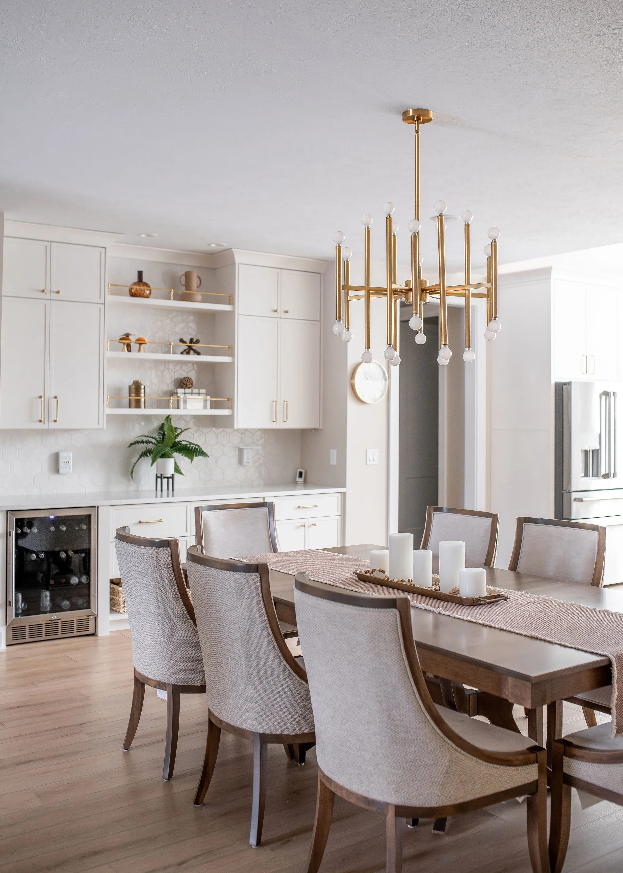 Elegant dining room with a wooden table, beige upholstered chairs, a modern gold chandelier, white kitchen cabinets, open shelves with decor, a green plant, and a wine fridge.