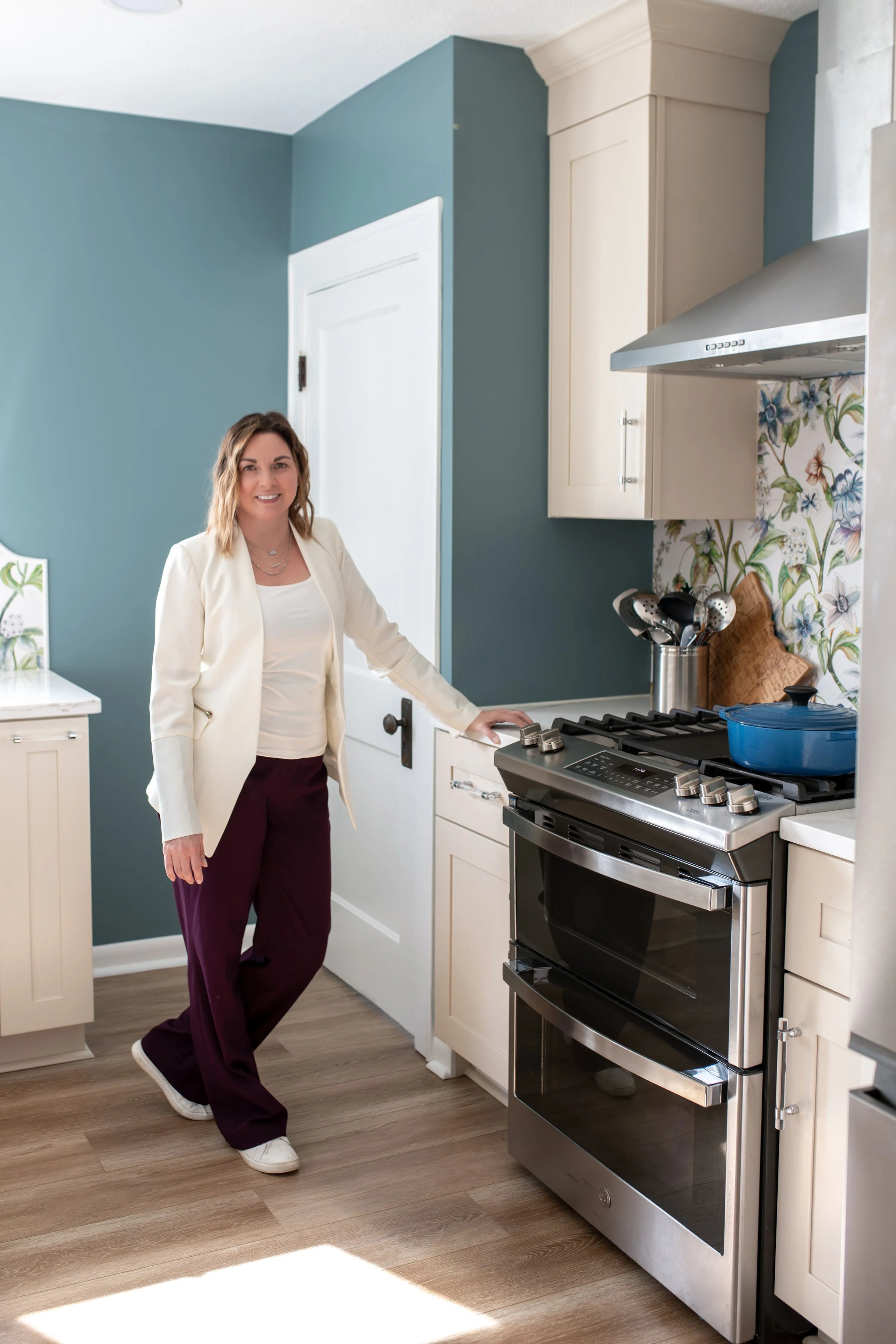 A woman in a cream blazer and burgundy pants standing in a kitchen with white cabinets, a blue wall, and a stainless steel stove.