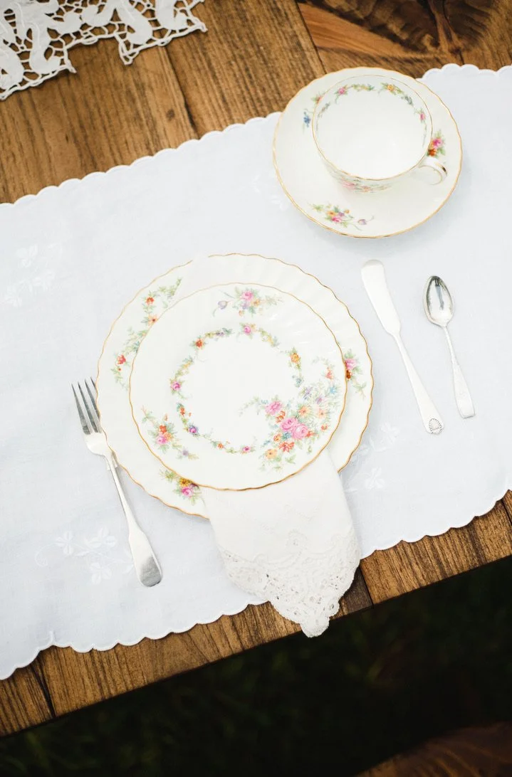 Elegant table setting with floral china plates, a teacup with matching saucer, a fork, a knife, and a spoon on a white embroidered tablecloth, placed on a wooden table.