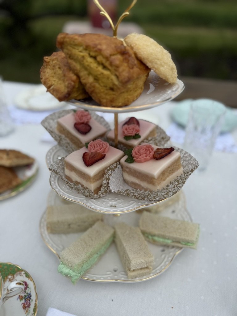Three-tiered dessert tray with savory scones on top, pink and white layered cakes with rose and strawberry decorations, and small sandwiches with various fillings.
