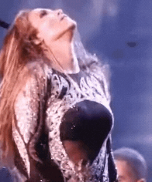 Person with long red hair with head tilted back, opening mouth wide, wearing a patterned black and grey top.