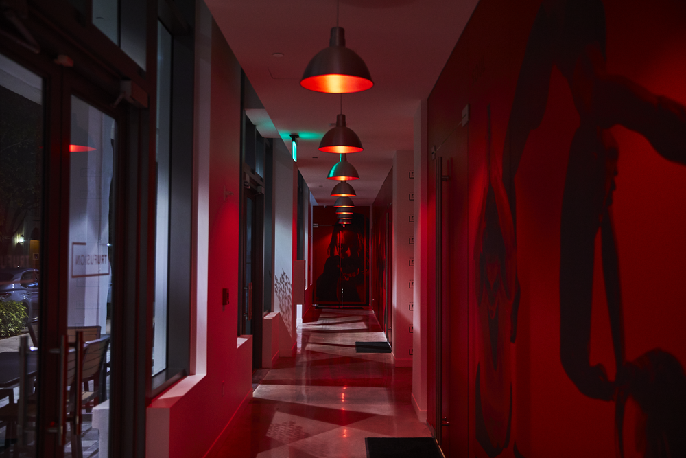 Dimly lit hallway with red and black wall art, black pendant lights, and reflections on a shiny floor, illuminated by red and teal lights.