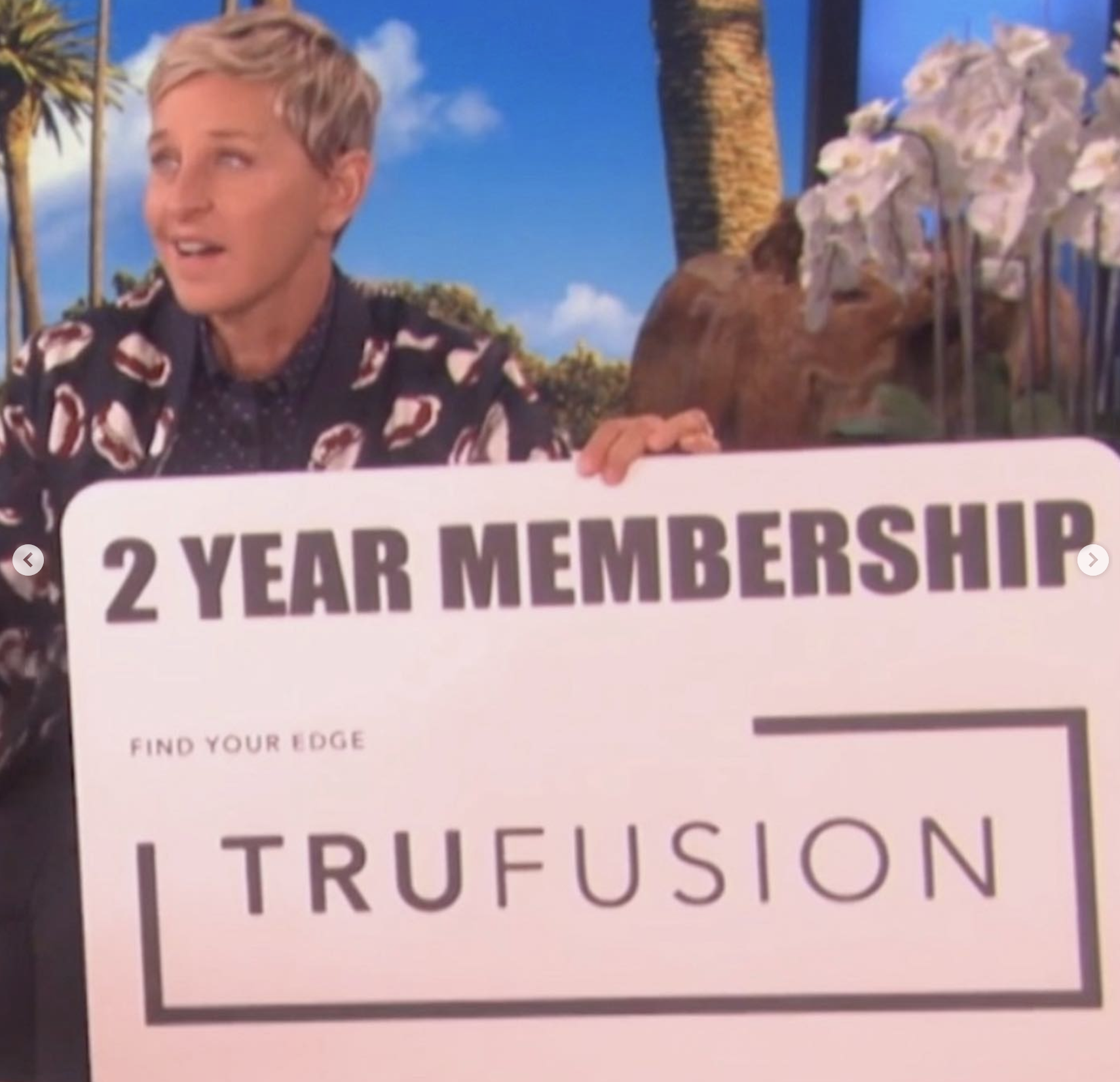 A person holding a sign advertising a 2-year membership for TruFusion, with a backdrop of tropical scenery including palm trees and a clear blue sky.