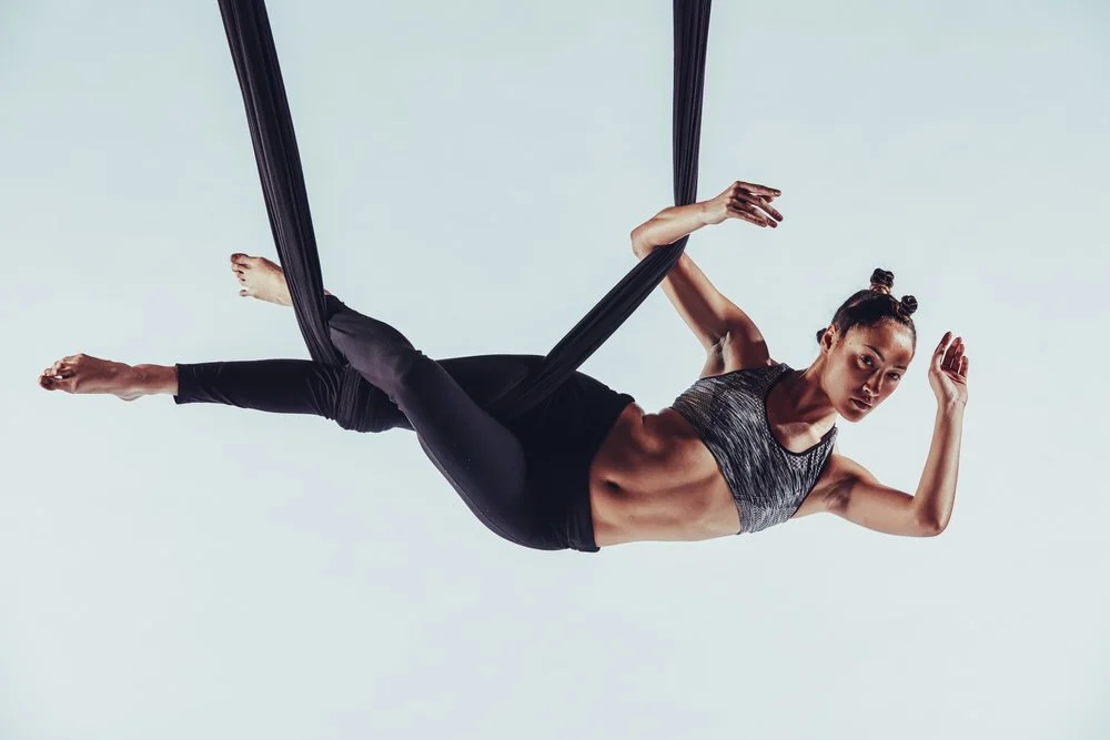 A woman performing aerial silk yoga or acrobatics, hanging horizontally in mid-air on black fabric against a plain light background.