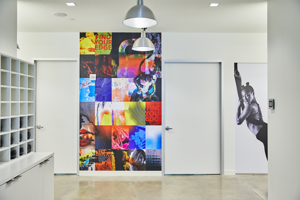Interior of a modern art studio or gallery with a colorful collage wall, a black-and-white photo of a woman stretching, and two silver pendant lamps hanging from the ceiling.