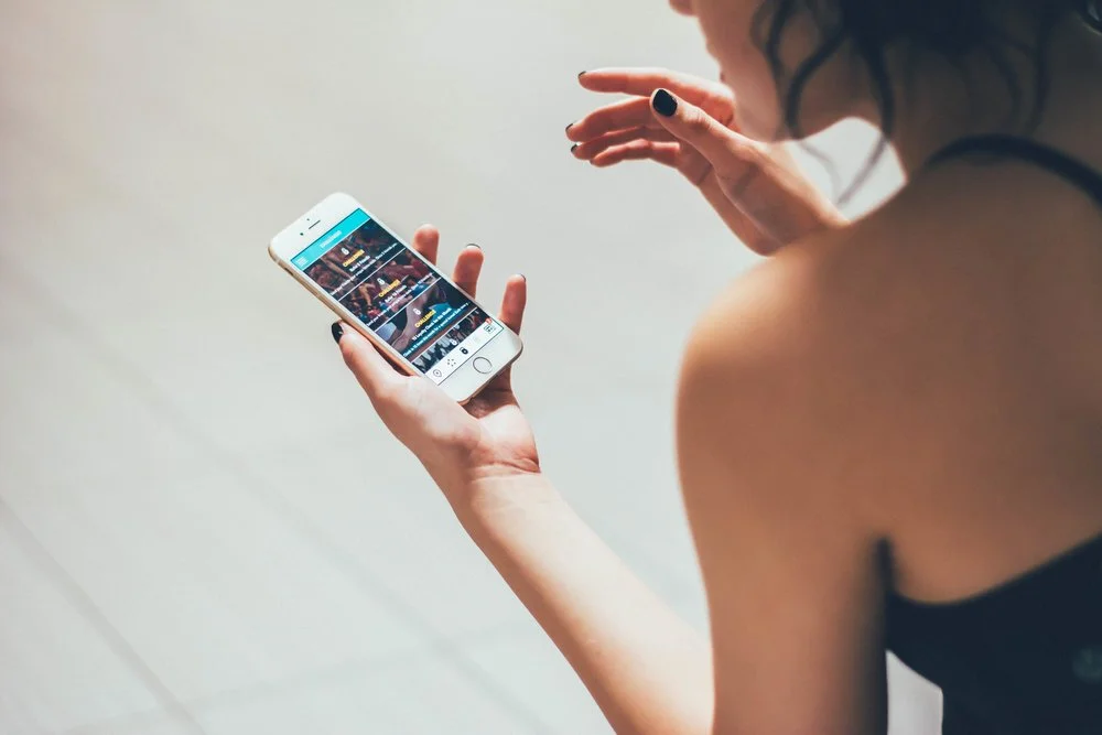 A woman with dark hair and nail polish is sitting and using a smartphone, browsing social media or a photo gallery.