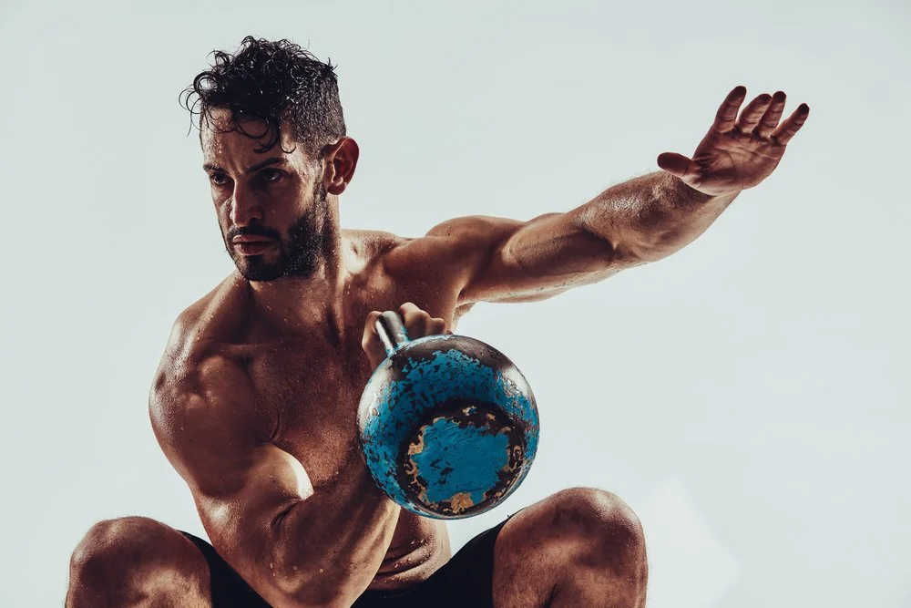 A muscular man with dark hair and a beard is holding a blue kettlebell while in a squatting position, with his left arm extended to the side.