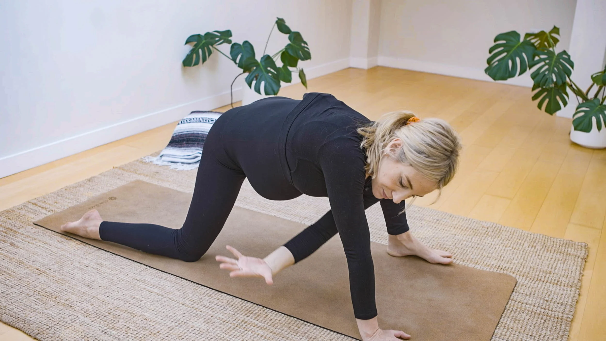 Pregnant woman practicing yoga on a brown mat indoors with large green plants in the background.