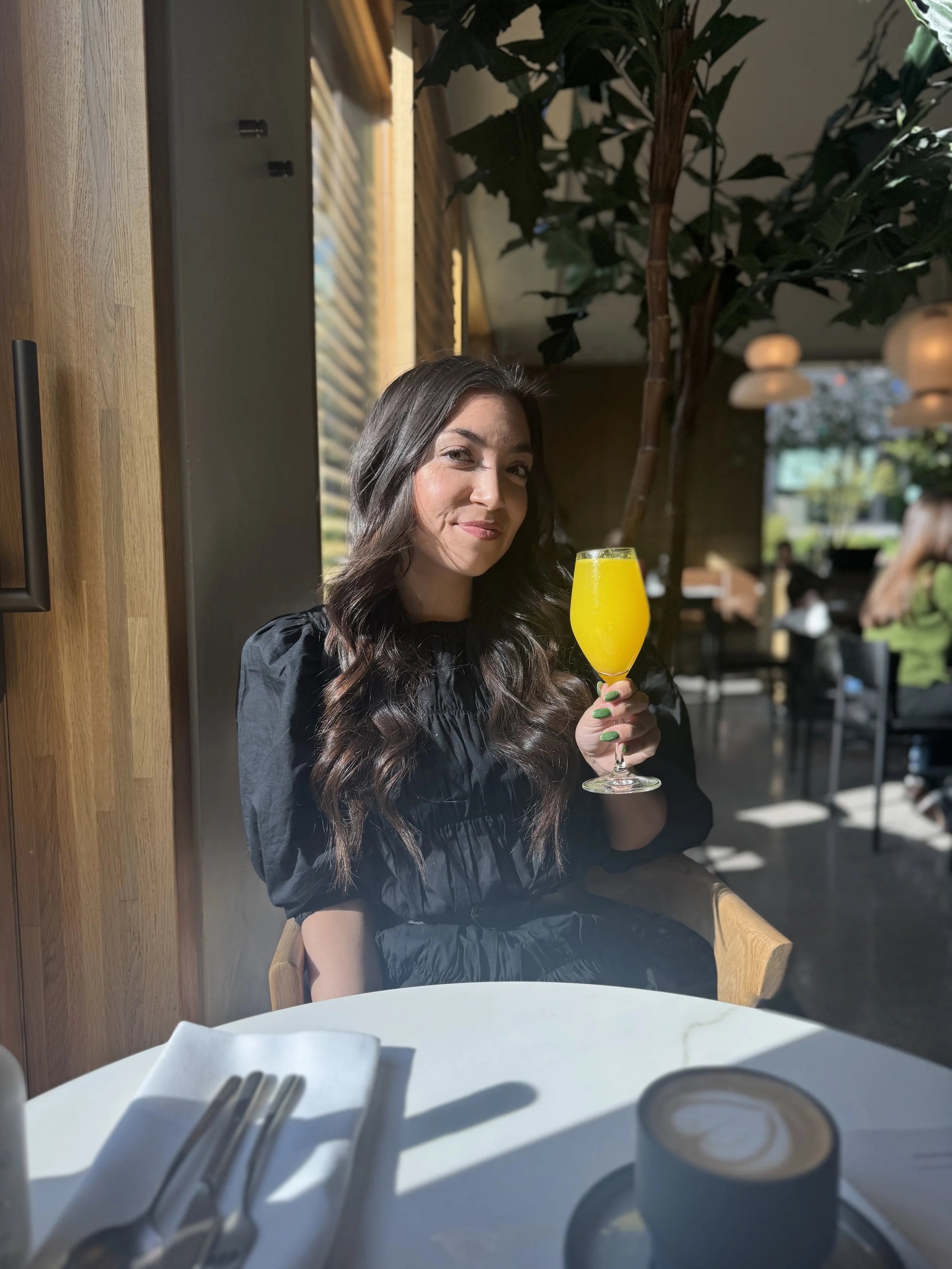 A woman with long dark wavy hair sitting at a table in a restaurant, holding a yellow cocktail glass, smiling at the camera.