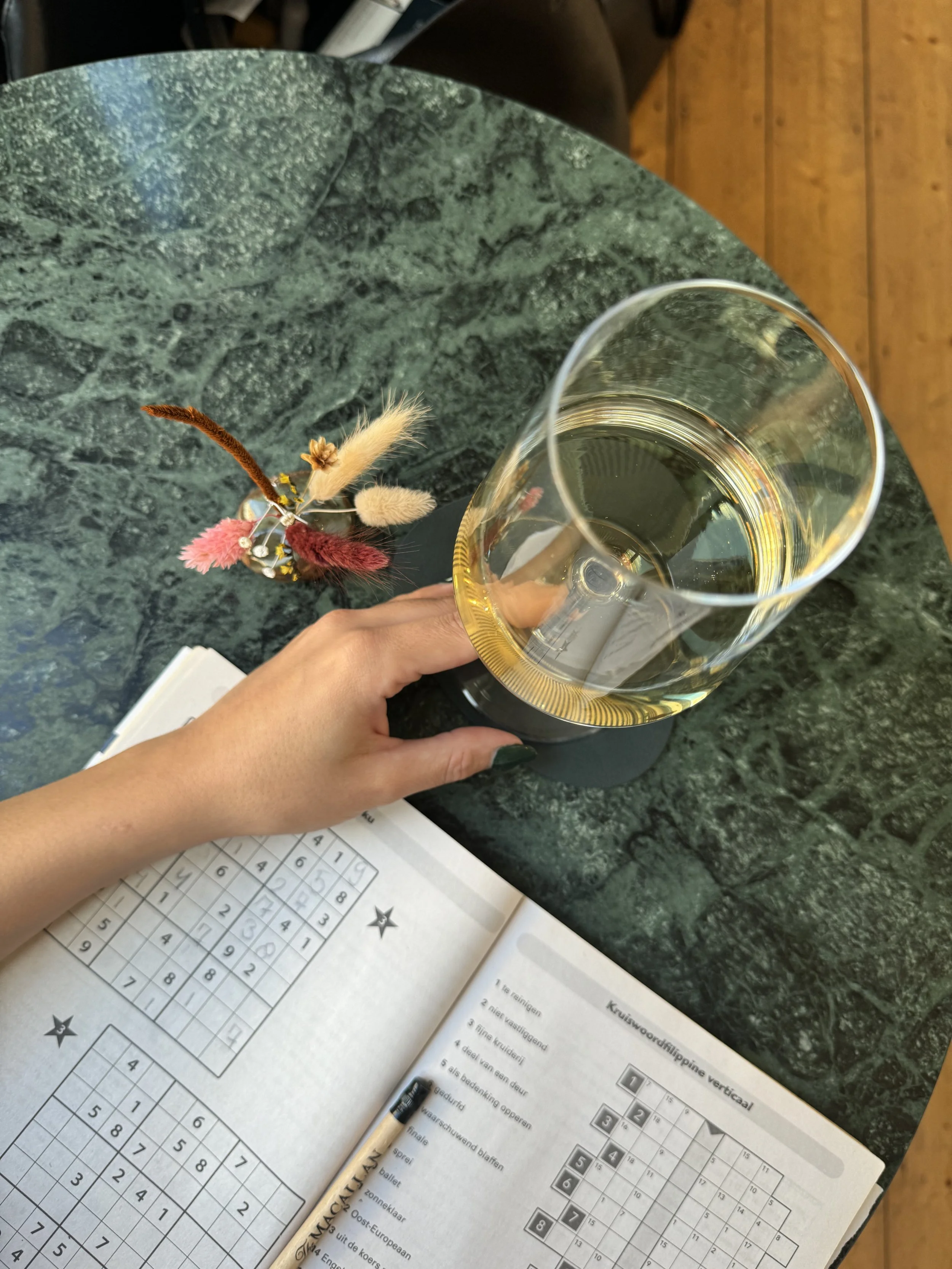 A person's hand holding a glass of white wine on a green marble table with a crossword puzzle book, a black pen, and a small colorful dried flower arrangement nearby.