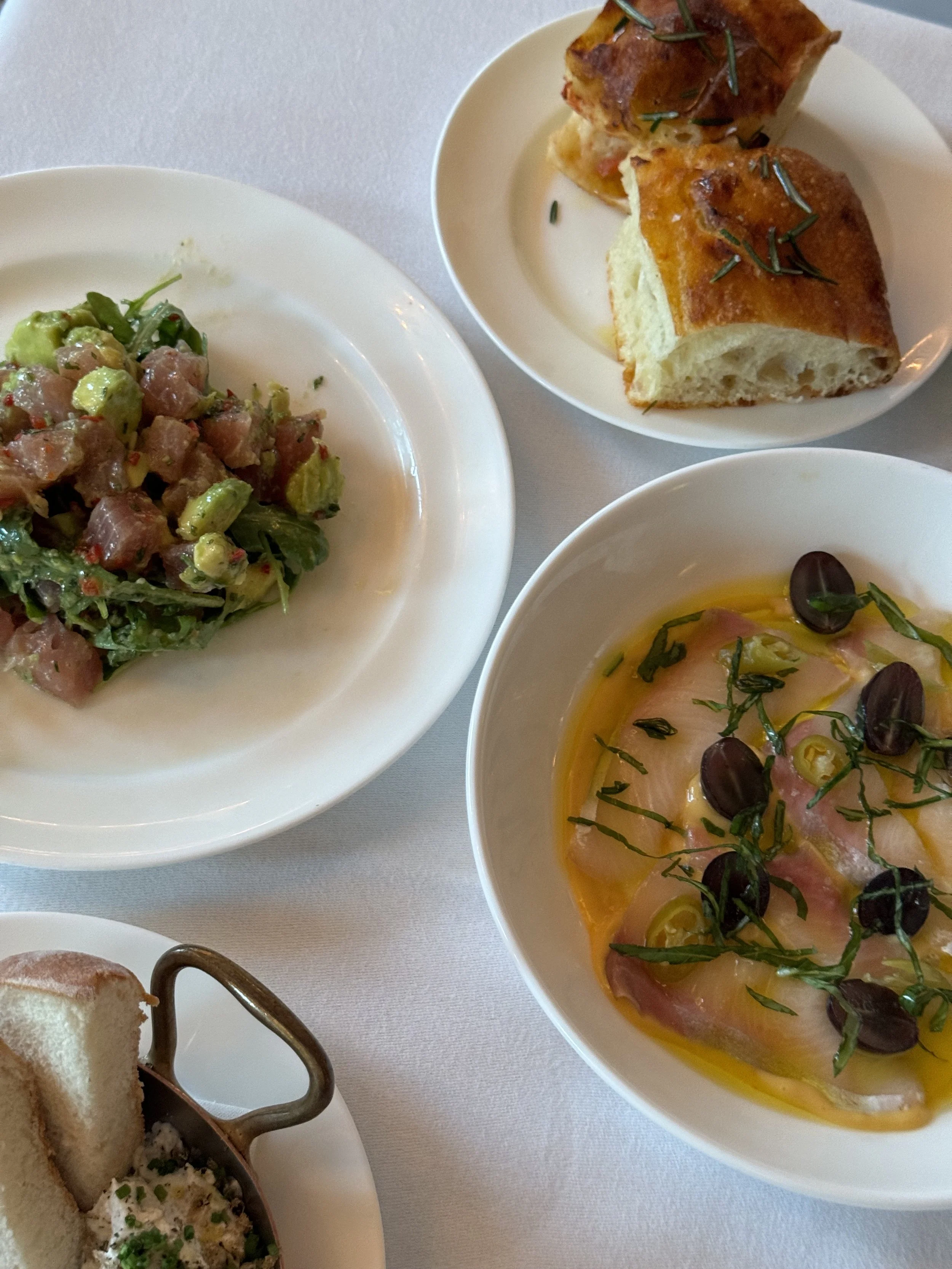 A table with four dishes: a small plate of chopped tuna and avocado salad, a bowl of cured fish with herbs and olives, a plate of bread with rosemary, and a dish of pâté with toast.