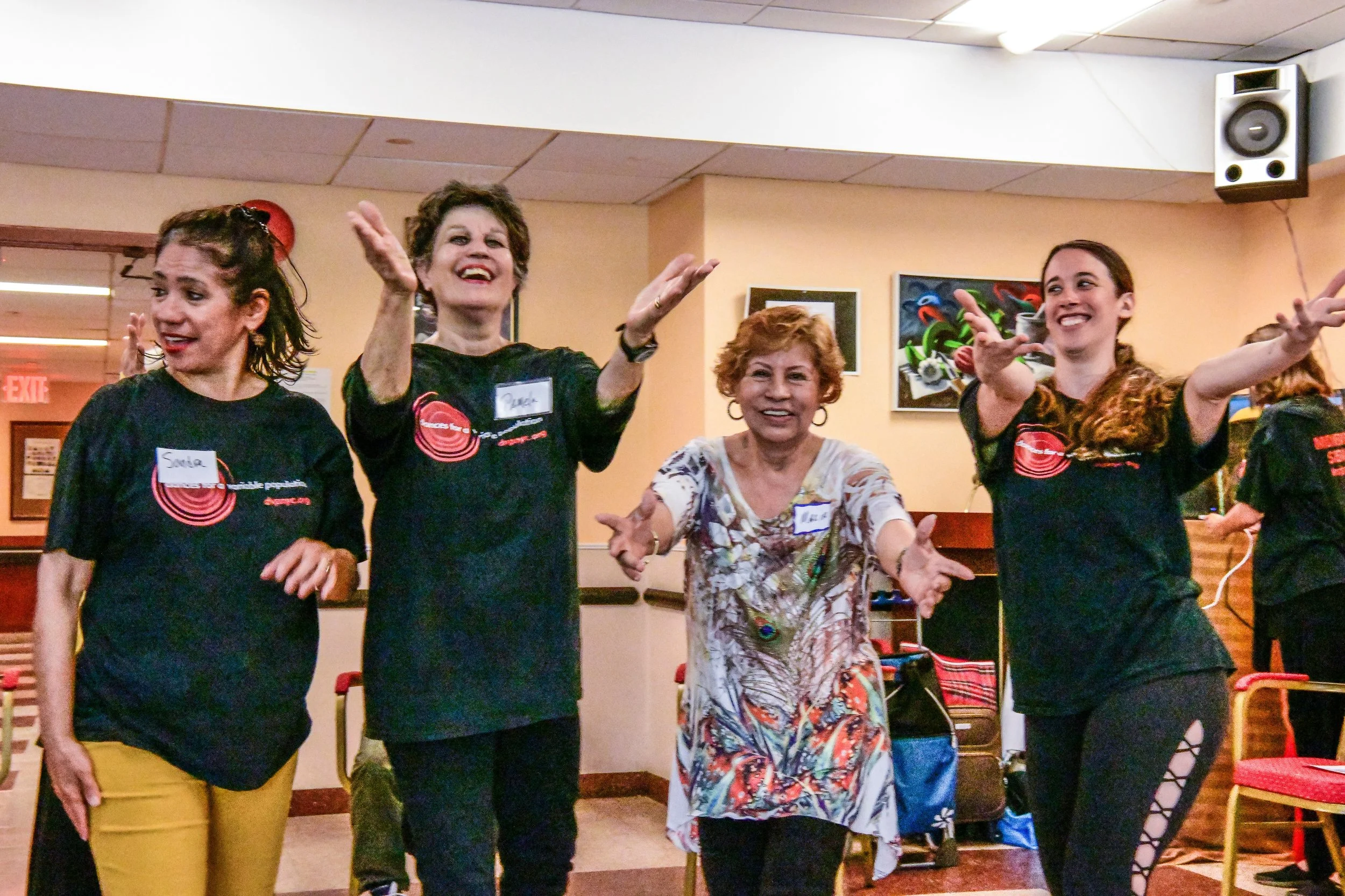 Group of women dancing and smiling indoors, wearing shirts and name tags.
