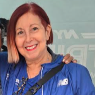 Smiling person with short black hair wearing a blue top, with greenery in the background.