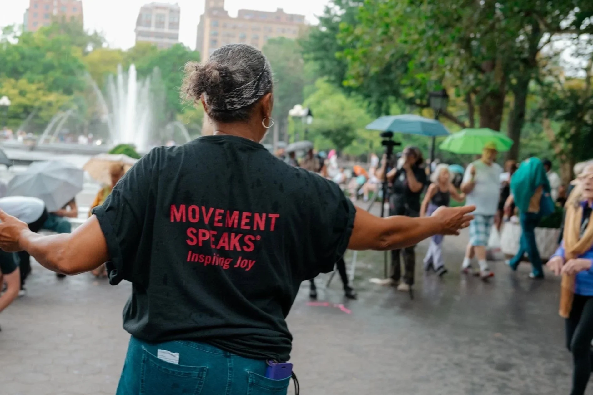 A person with short hair, wearing a black t-shirt with red text that reads "MOVEMENT SPEAKS Inspiring Joy," gesturing with arms outstretched in a park with people holding umbrellas, trees, and high-rise buildings in the background.