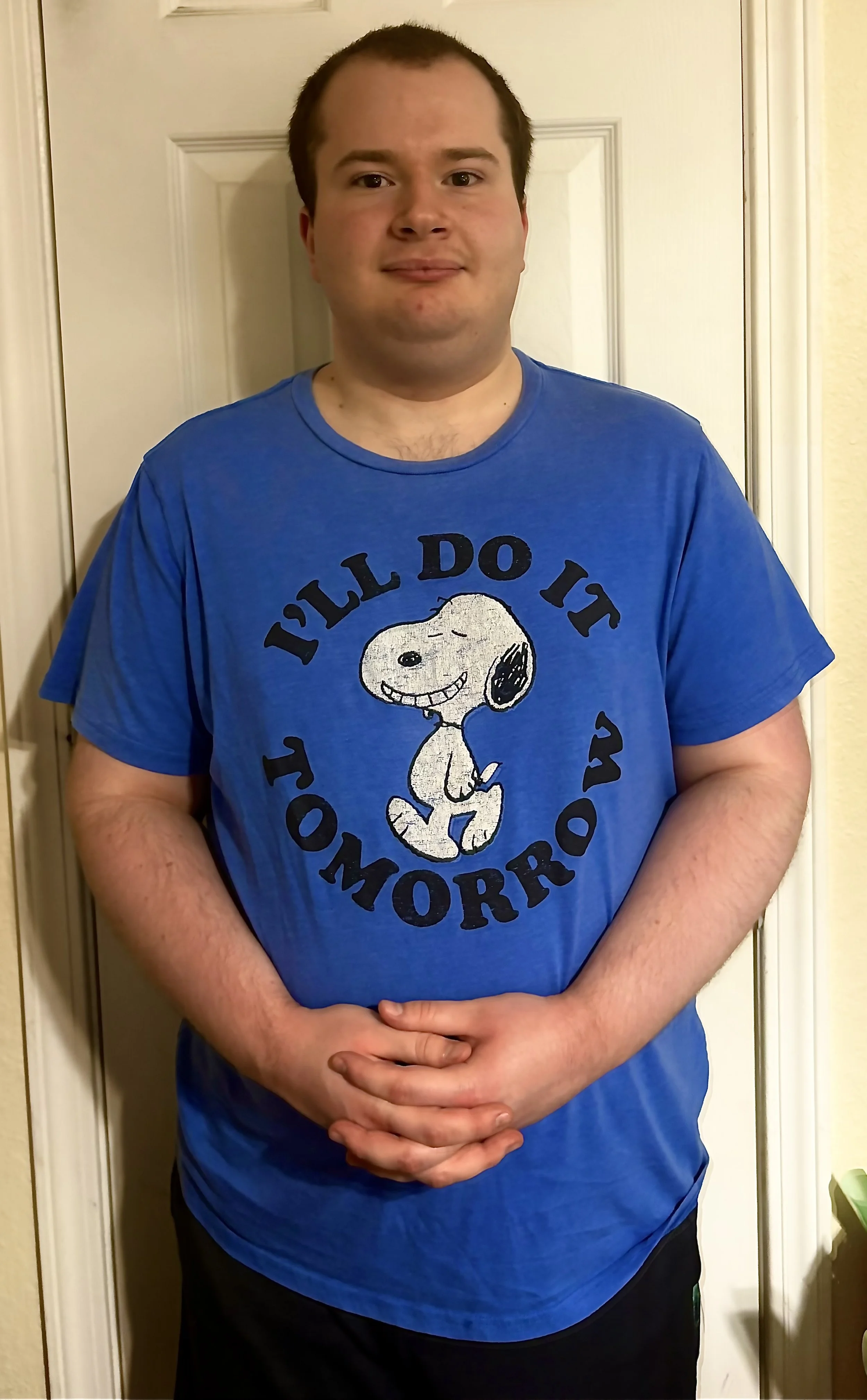A man with short brown hair and light skin stands indoors in front of a white door, wearing a blue T-shirt featuring a cartoon character from Peanuts, Snoopy, with the text 'It'll Do It Tomorrow' surrounding the character. His hands are clasped in front of him.