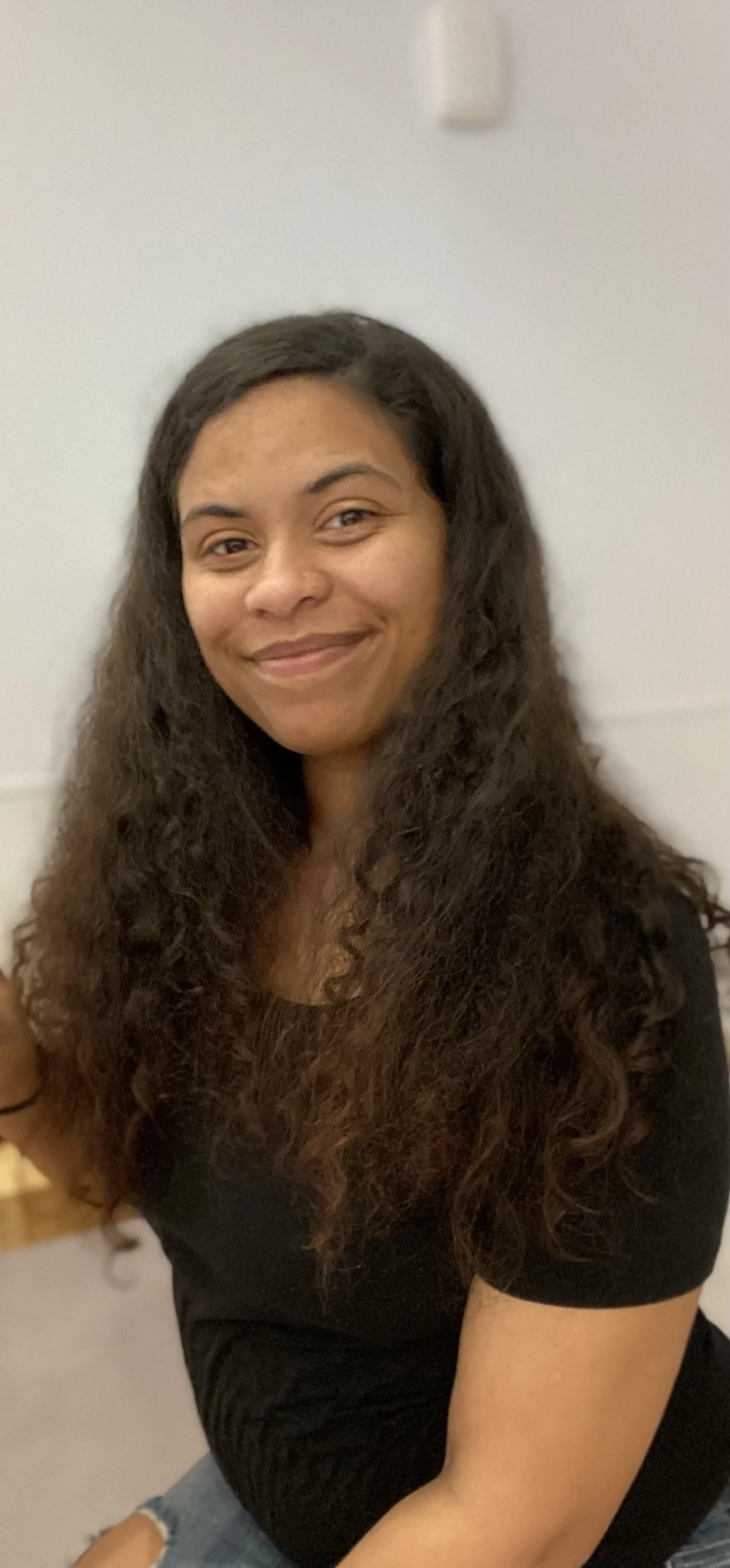 A young woman with long, curly brown hair sitting in a room with white walls, smiling at the camera.