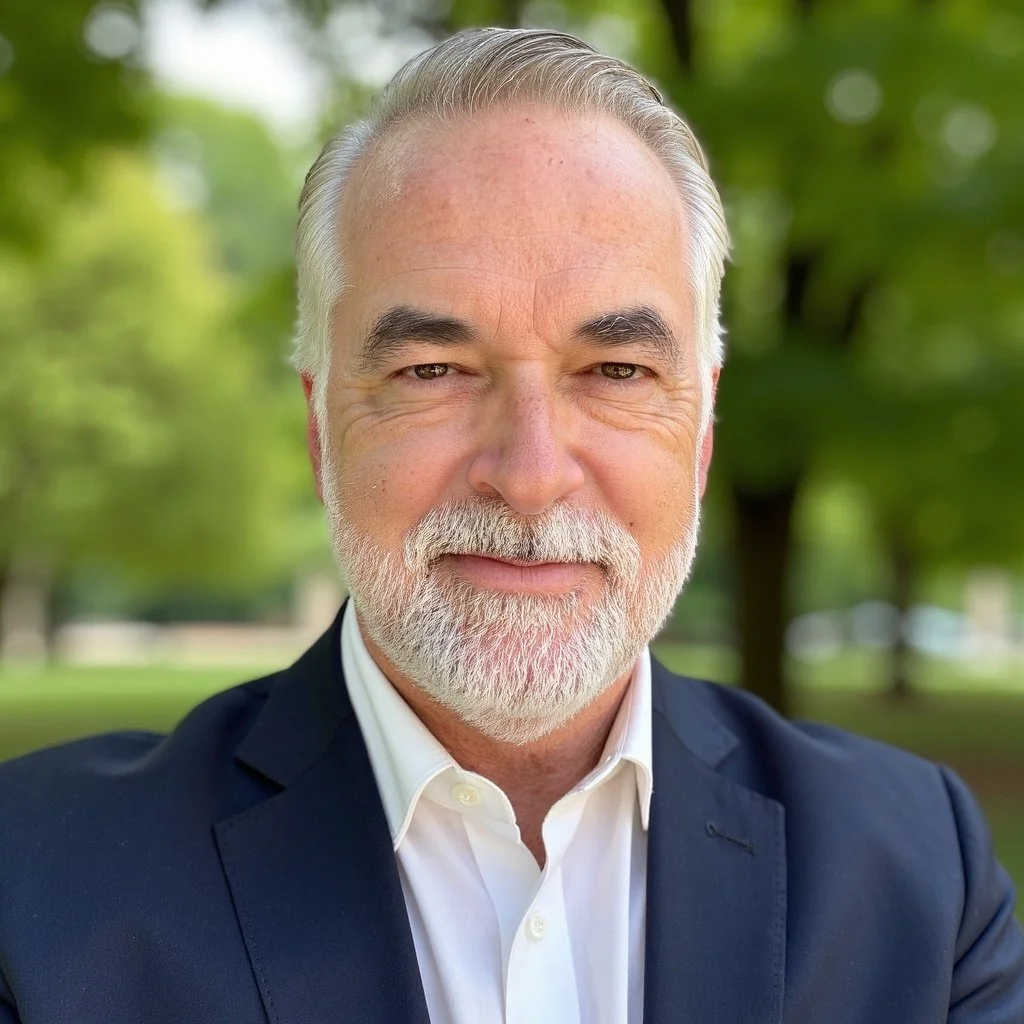A close-up portrait of an older man with gray hair, a full beard, and mustache, wearing a dark suit and white shirt, outdoors with green trees in the background.