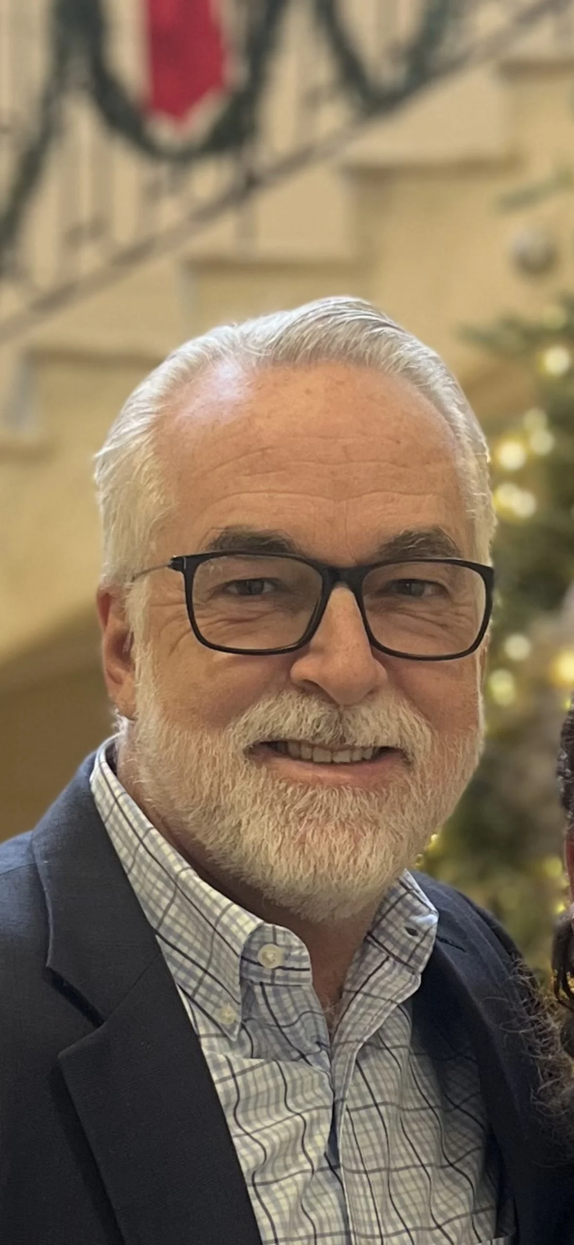 Middle Aged grey haired man with black glasses smiling with a grey plaid shirt collar and a blurred background