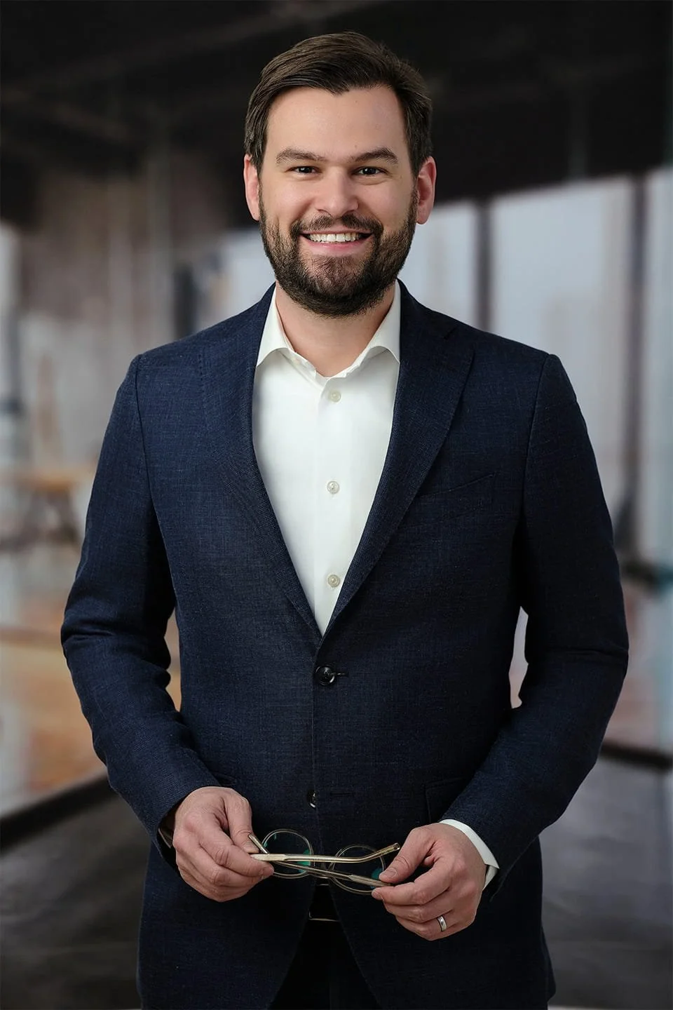 A man in a navy suit holding glasses, smiling in an indoor setting.
