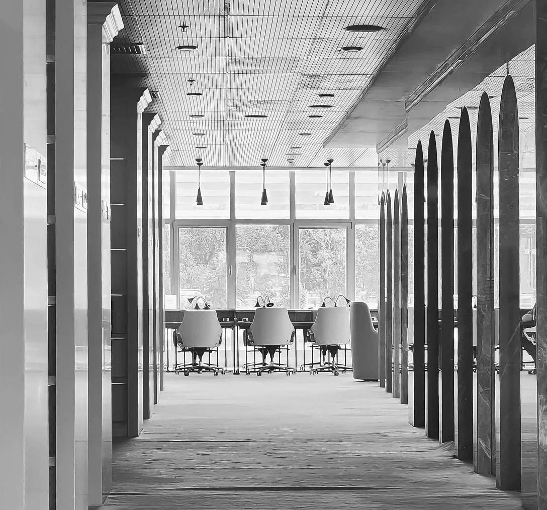 Empty modern office space with chairs arranged around a table near large windows, viewed through a corridor with vertical wooden dividers.