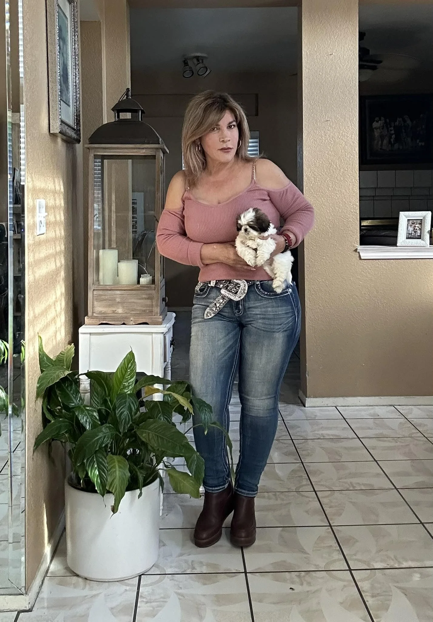A woman holding a small puppy in a home interior, standing next to a potted plant and a small white table with candles on it.