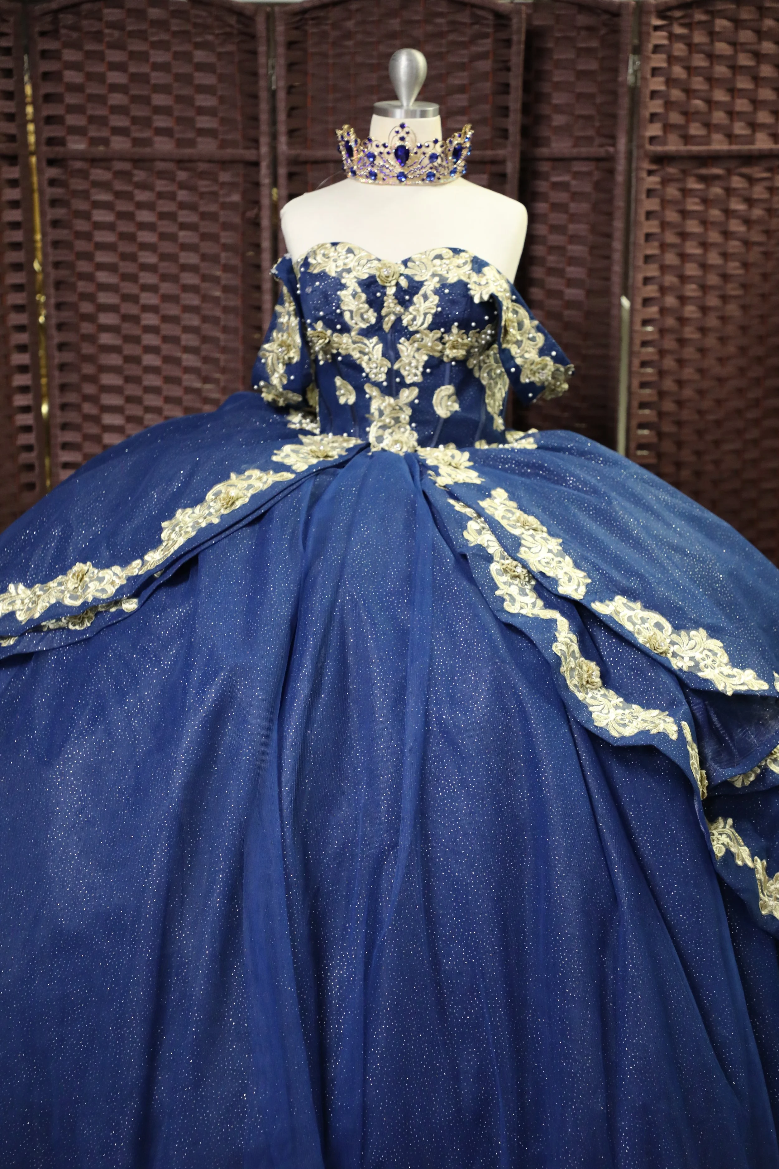 A royal blue ball gown with gold and white embroidery, displayed on a mannequin with a matching crown necklace and a silver ornamental top, against a wooden backdrop.