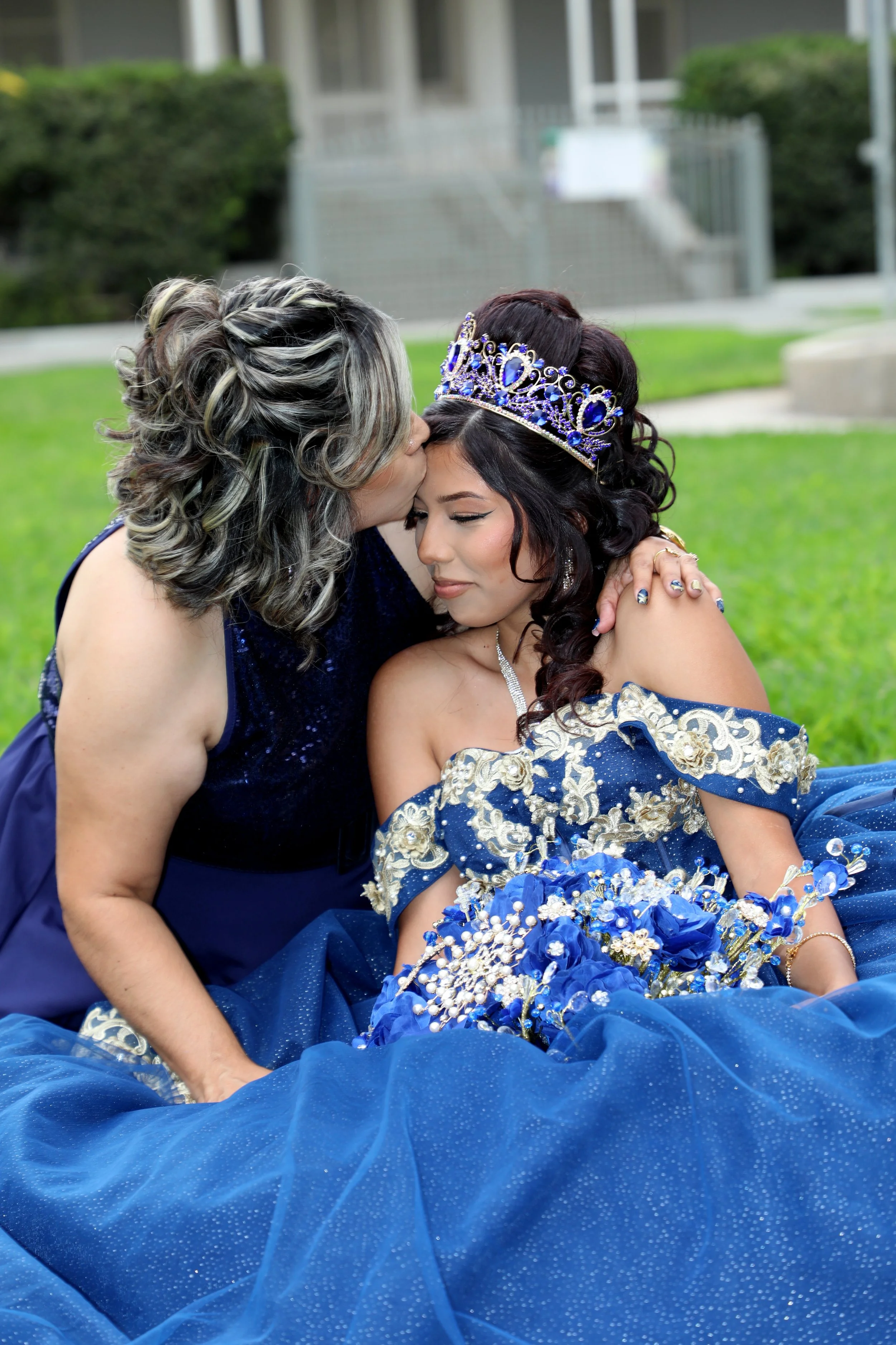 A young woman dressed in a blue princess gown with gold embroidery and a tiara on her head sitting on grass, while an older woman, possibly her mother, kisses her forehead in a loving gesture during a celebration or special occasion.