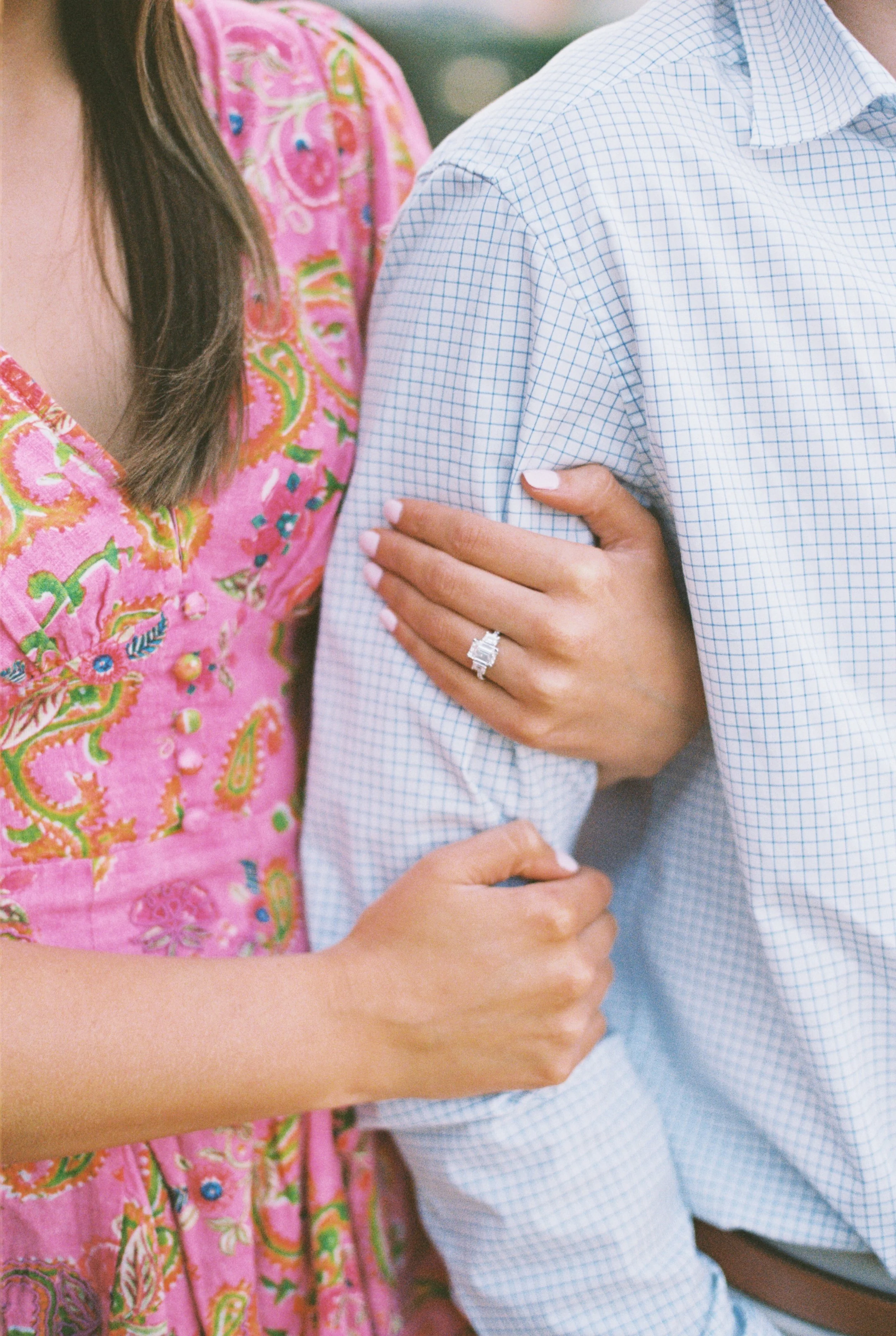 Close-up of a woman's arm linked with a man's arm; she is wearing a pink dress with colorful patterns and a diamond engagement ring.
