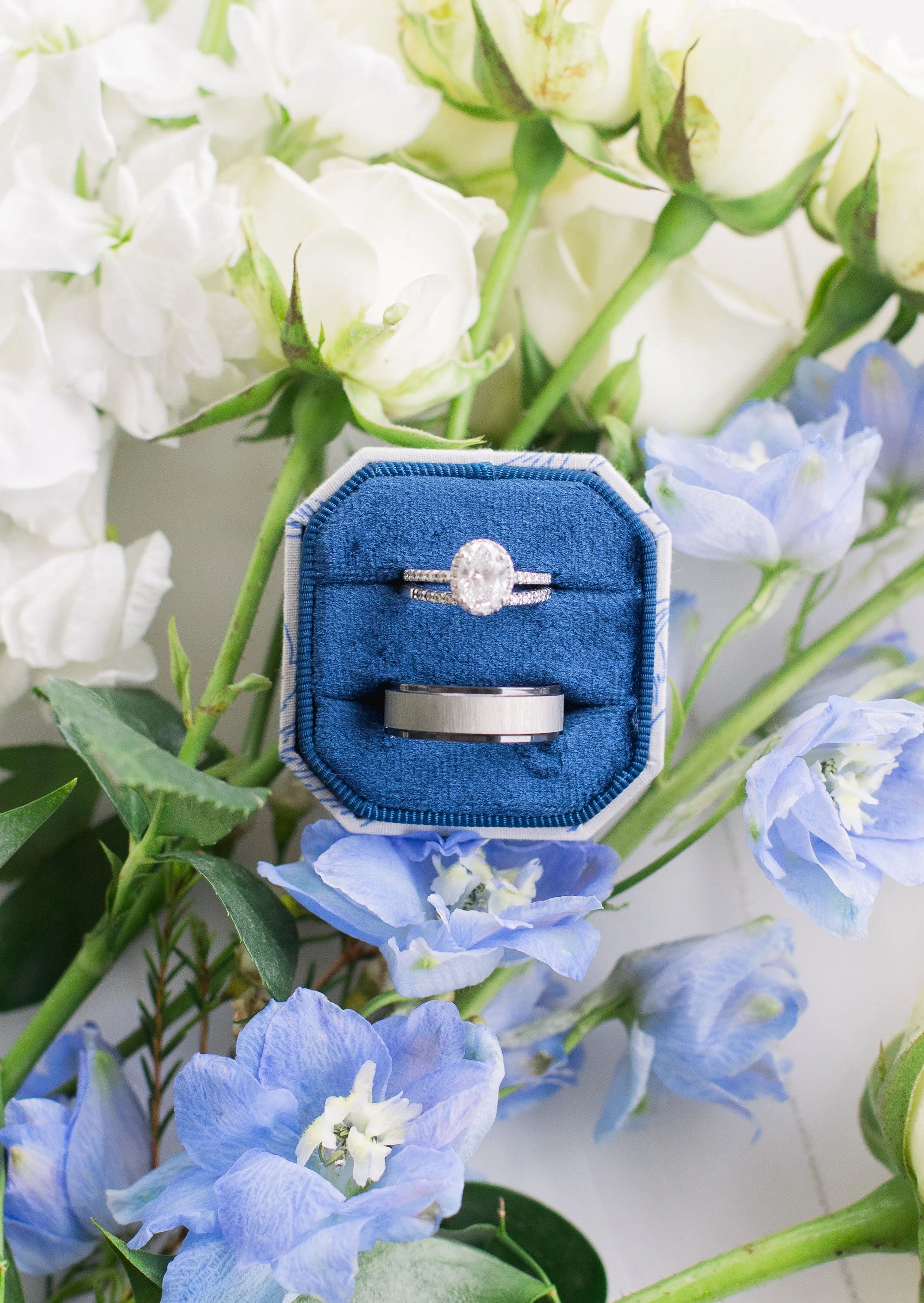 Wedding rings in a blue velvet box surrounded by white and blue flowers.