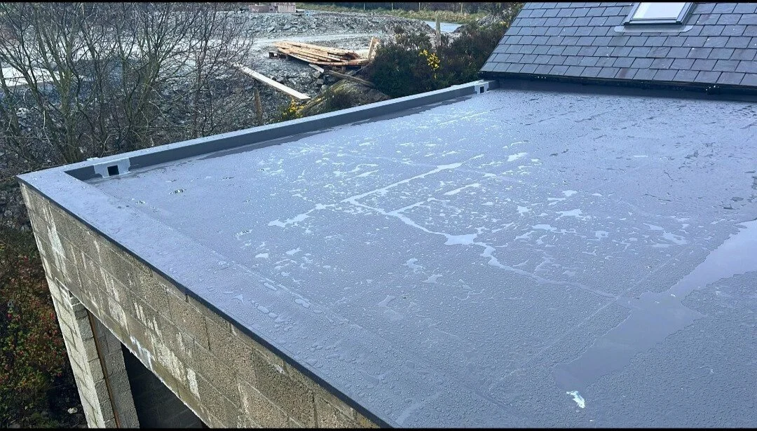 Flat roof with water droplets on its surface and a neighboring roof with shingles, trees, and construction debris in the background.