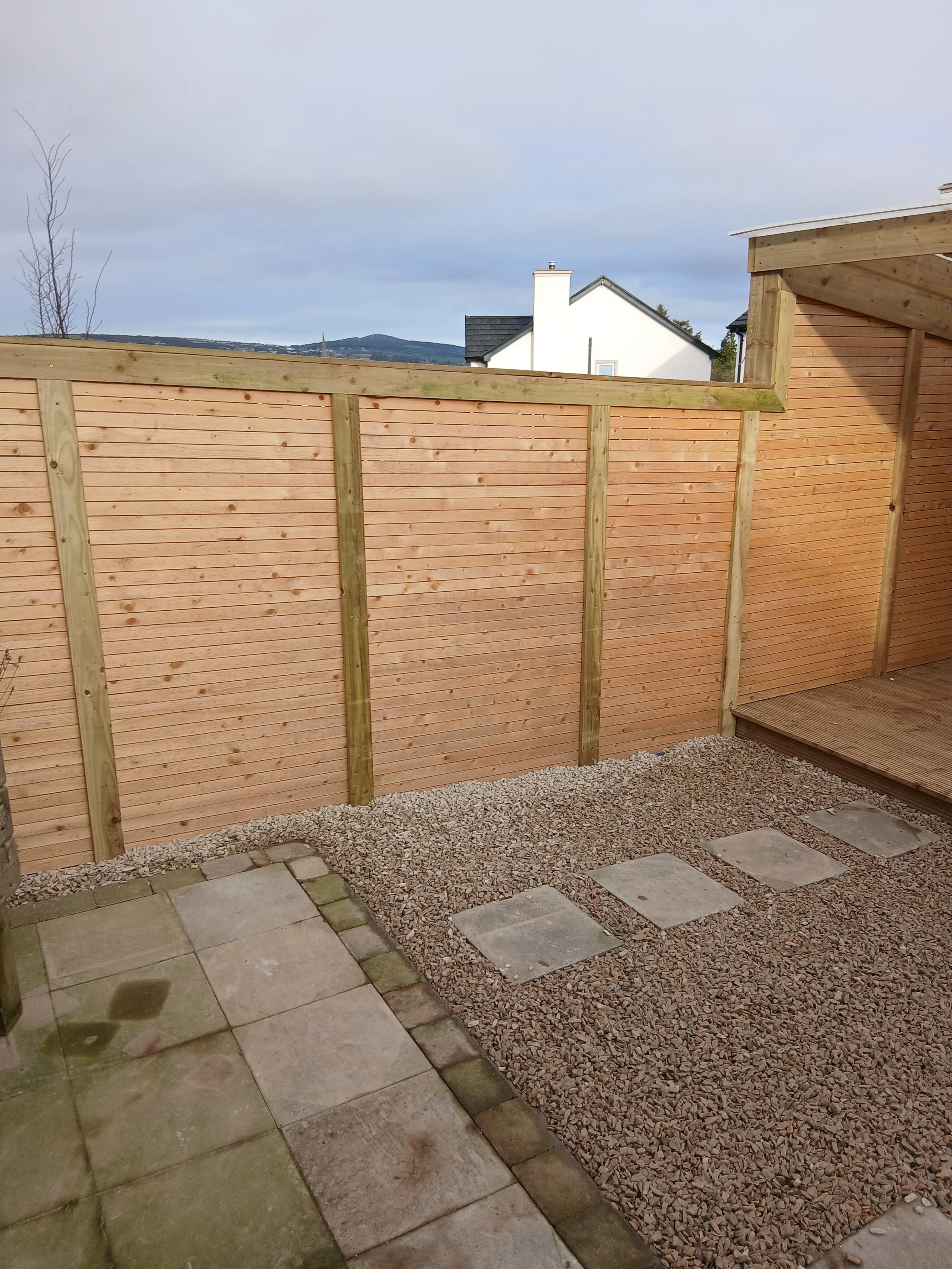 A backyard patio with a sitting area of stone and concrete tiles. Behind a newly built tan wooden fence with vertical planks and greenish posts, there is an open gravel area with square stepping stones. In the background, rooftops and a cloudy sky are visible.