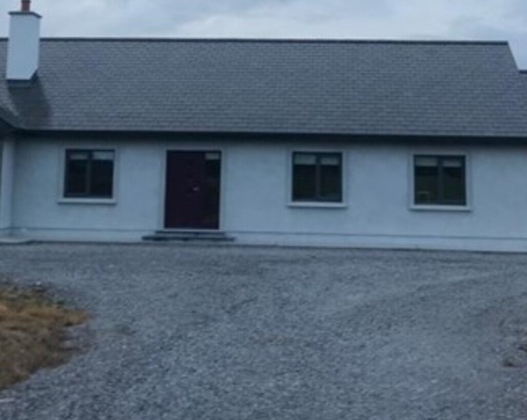 Front view of a house with a gray facade, three windows, a red front door, and a gravel driveway.