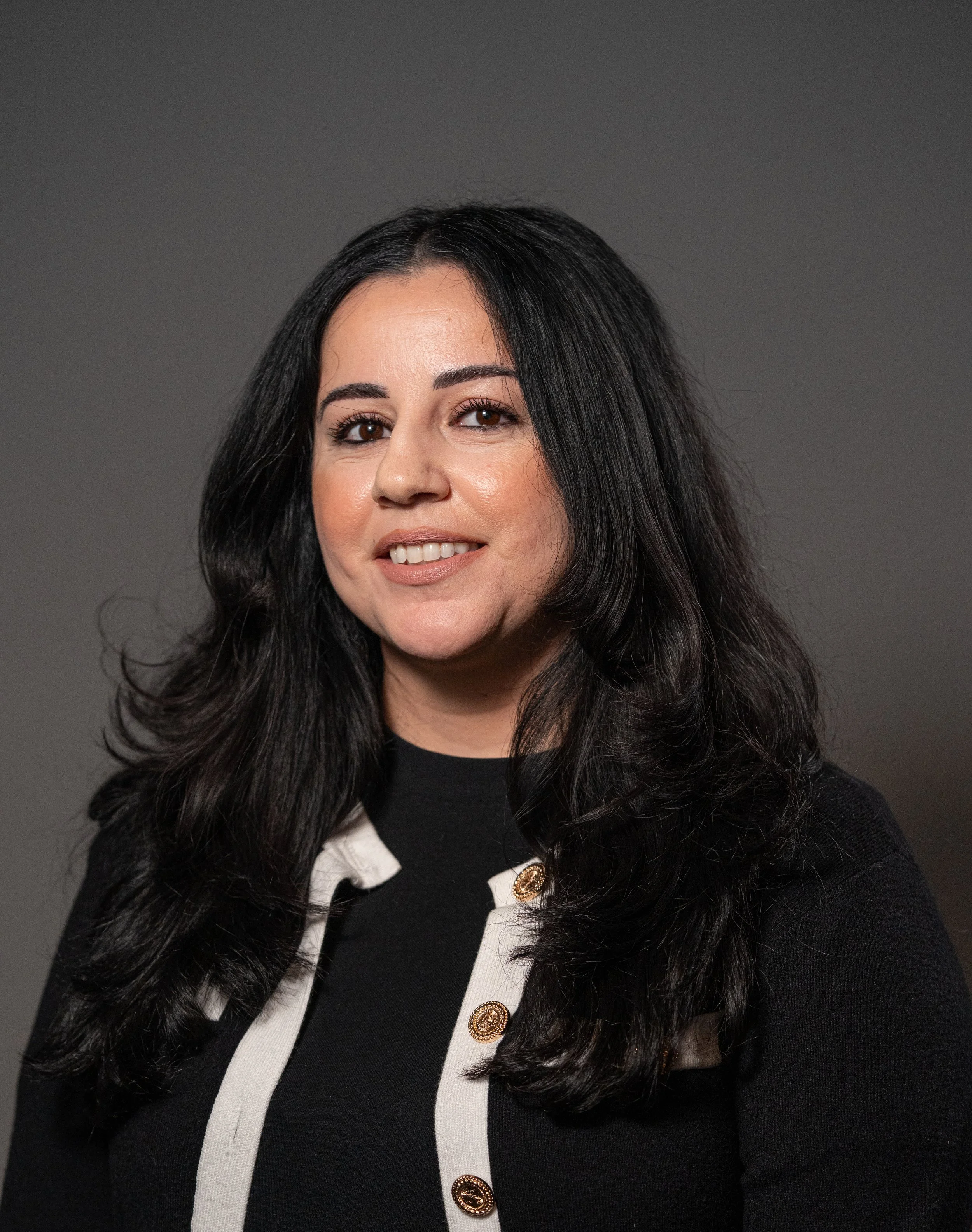 Portrait of a woman with long black hair, wearing a black top with white accents and gold buttons, smiling against a gray background.