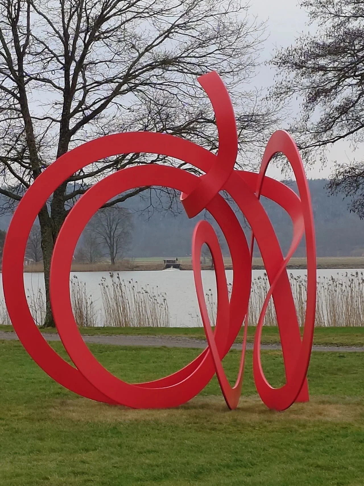 En stor, röd skulptur formad som en dekorativ och abstrakt design placerad på en gräsyta nära en sjö, med träd och berg i bakgrunden.