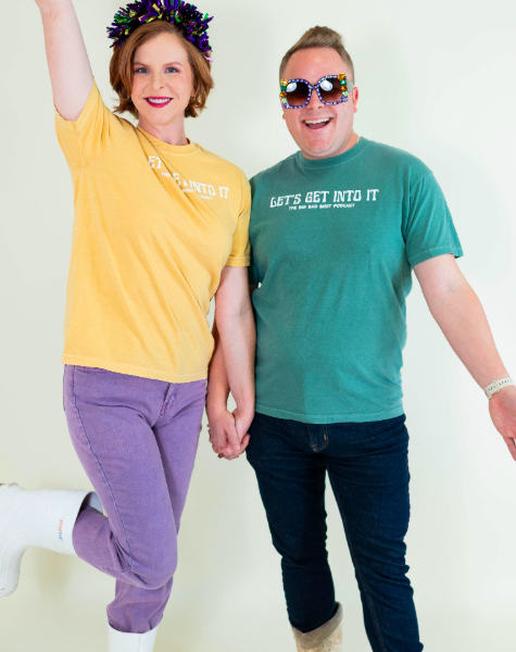 Two people celebrating, holding hands, smiling, wearing fun glasses and colorful clothing, standing against a plain background.