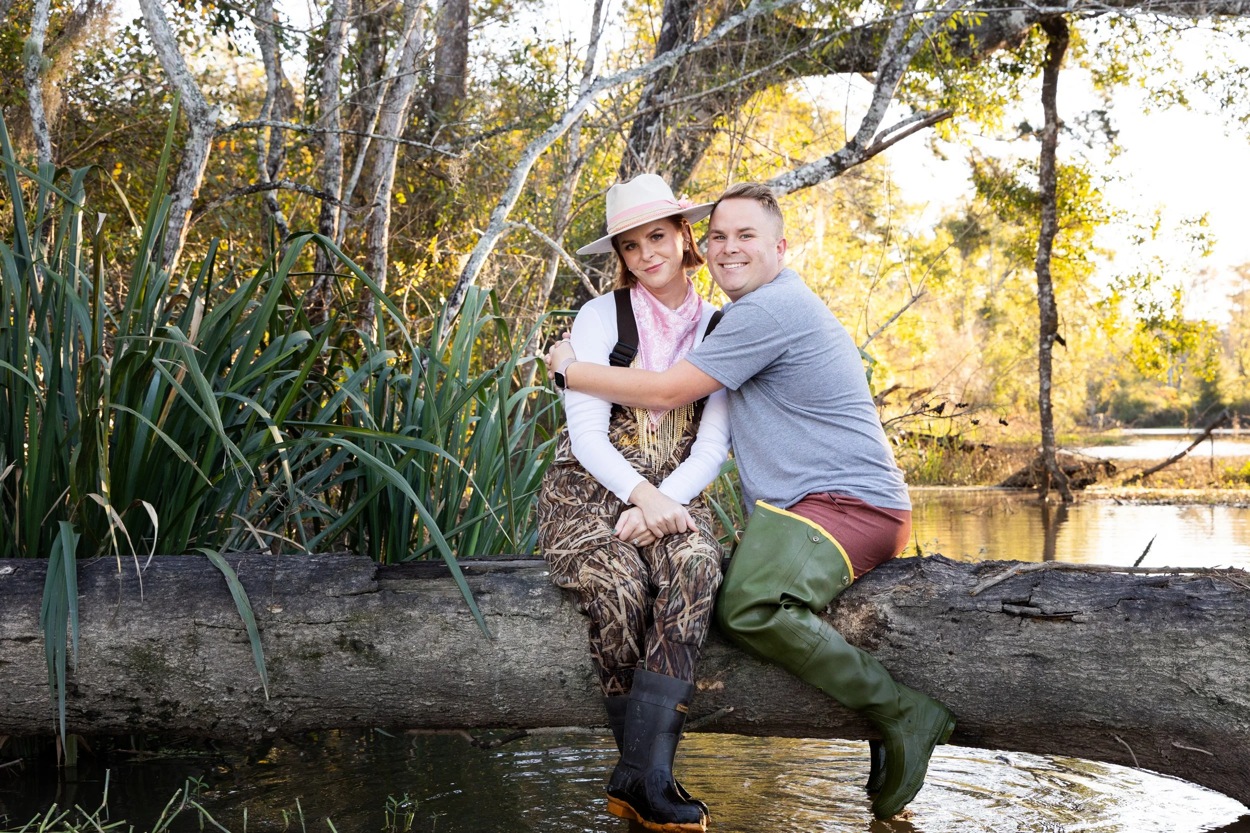Two people, a woman and a man, sitting on a fallen tree over a body of water in a wooded area during autumn, smiling and hugging.