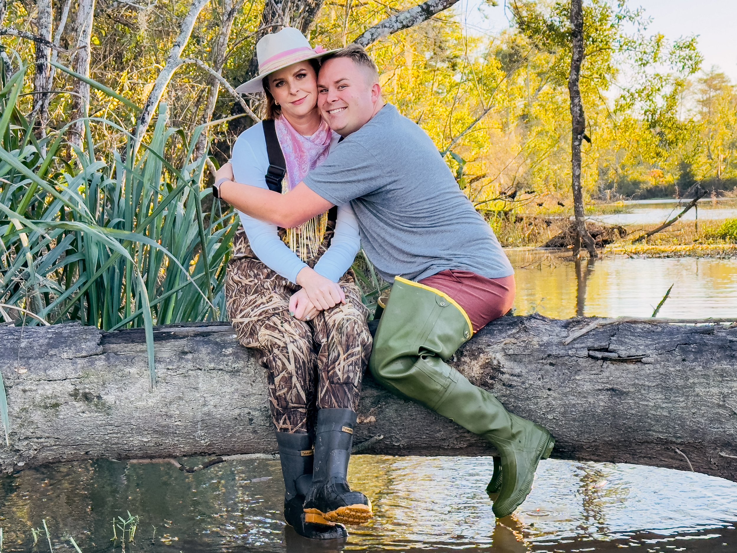 A woman and a man sitting together on a tree trunk over water in a natural setting with trees and yellow foliage, smiling and hugging.
