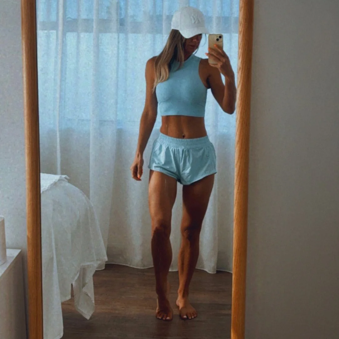 Woman taking a mirror selfie, wearing a light blue sports bra, matching shorts, a white cap, and barefoot in a bedroom with white curtains and wooden flooring.