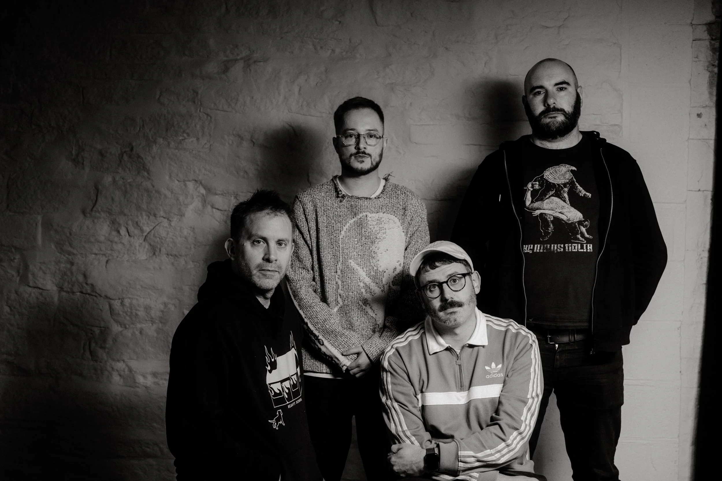 Black and white photo of five men with various hairstyles and glasses, posed against a rough-textured wall.