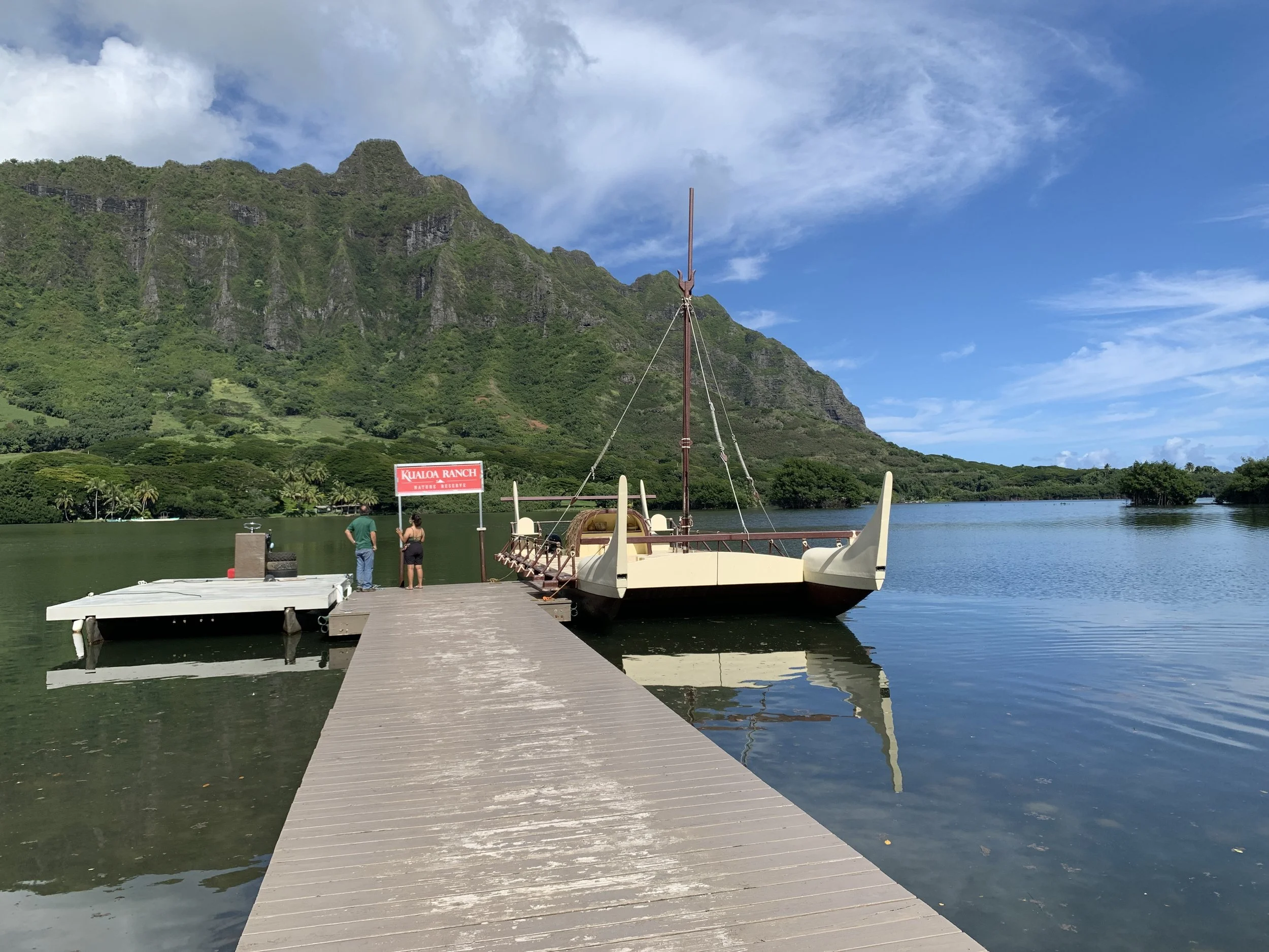 Scenic view of a traditional boat docked near a wooden pier, with a mountain and lush greenery in the background at Kualoa Ranch, Hawaii. Hawaii film, video, photo production service
