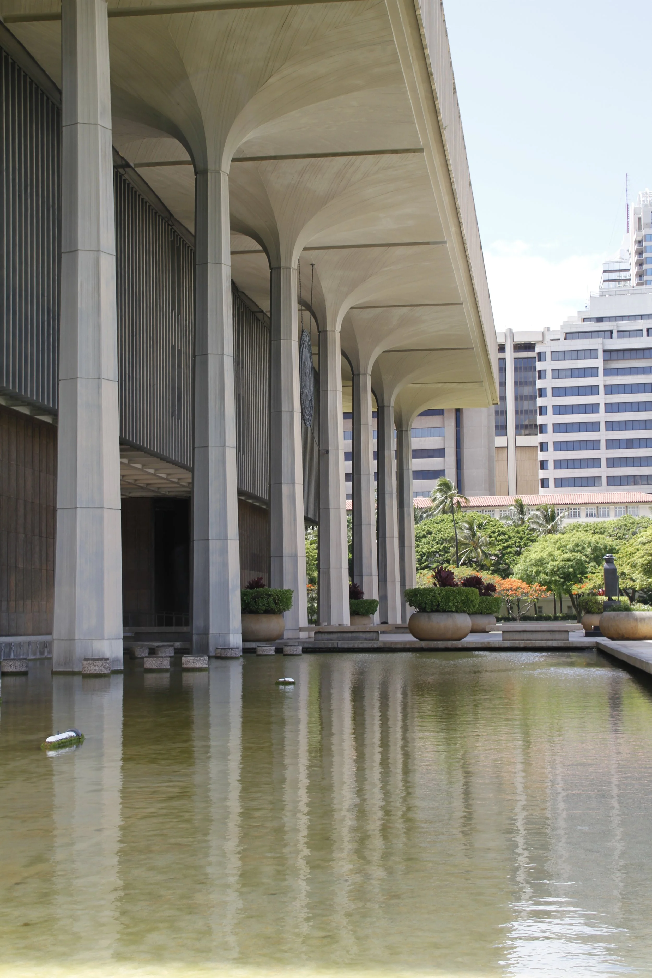Modern architectural building with large columns and a reflecting pool, surrounded by greenery and adjacent to an urban skyline. Hawaii film, video, photo production service