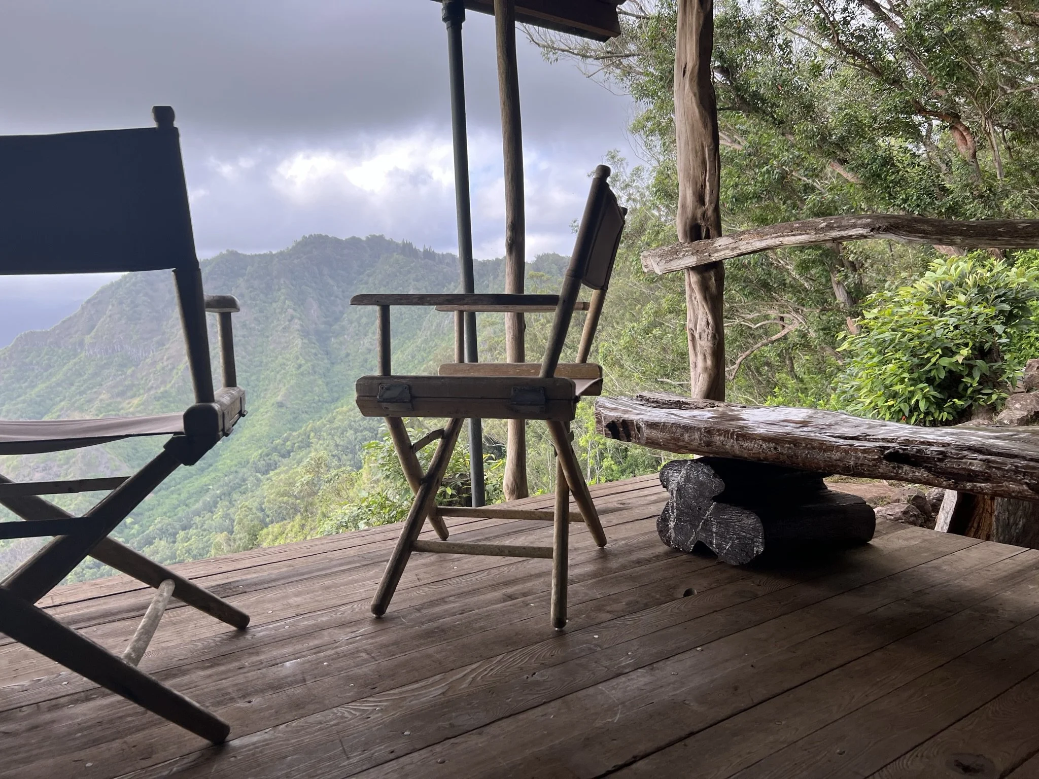 Wooden deck with chairs overlooking a lush green valley and mountains, surrounded by forest and cloudy sky. Hawaii film, video, photo production service