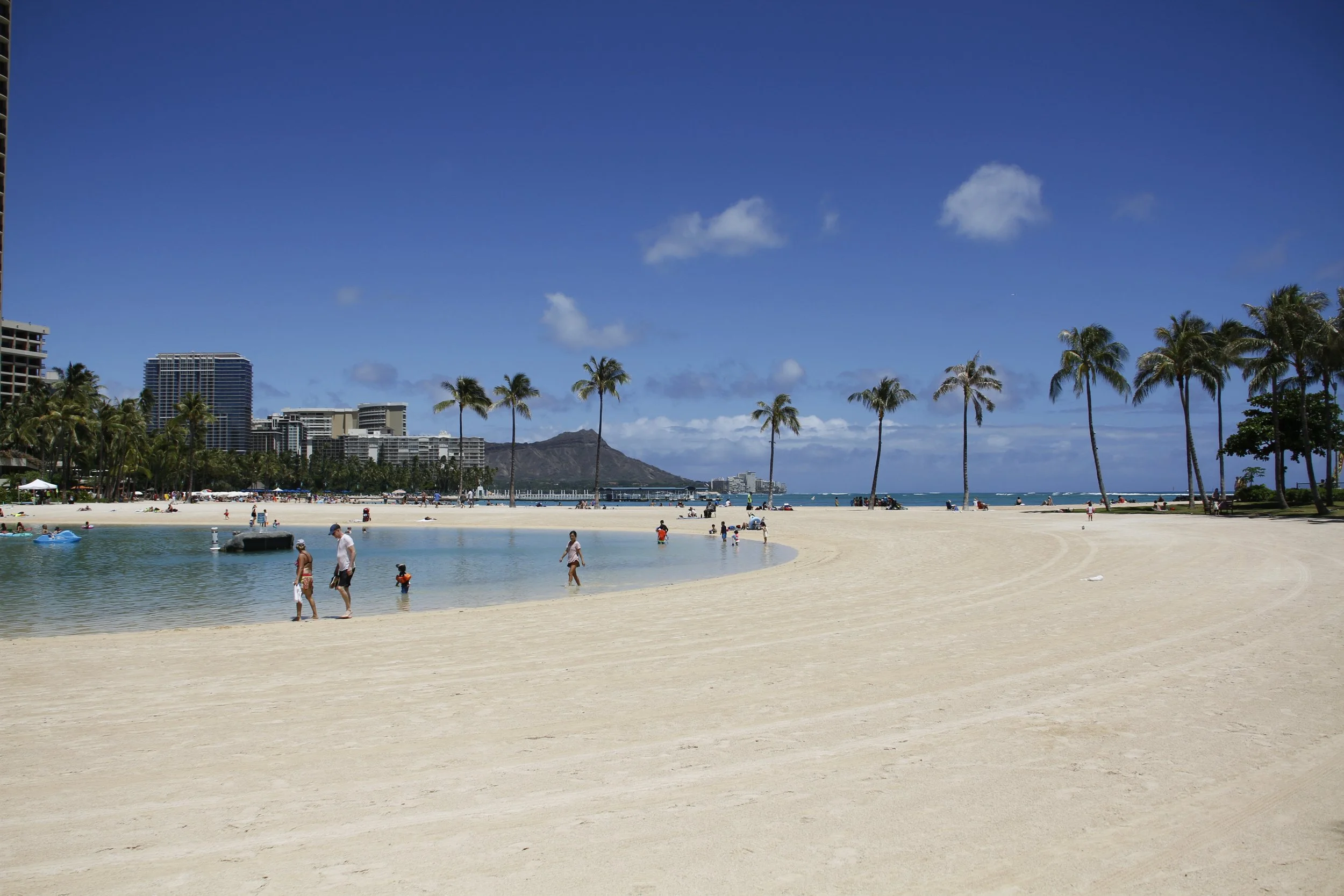 People enjoying a sunny day at a sandy beach with palm trees, a large pool, and distant mountains in the background. Hawaii film, video, photo production service
