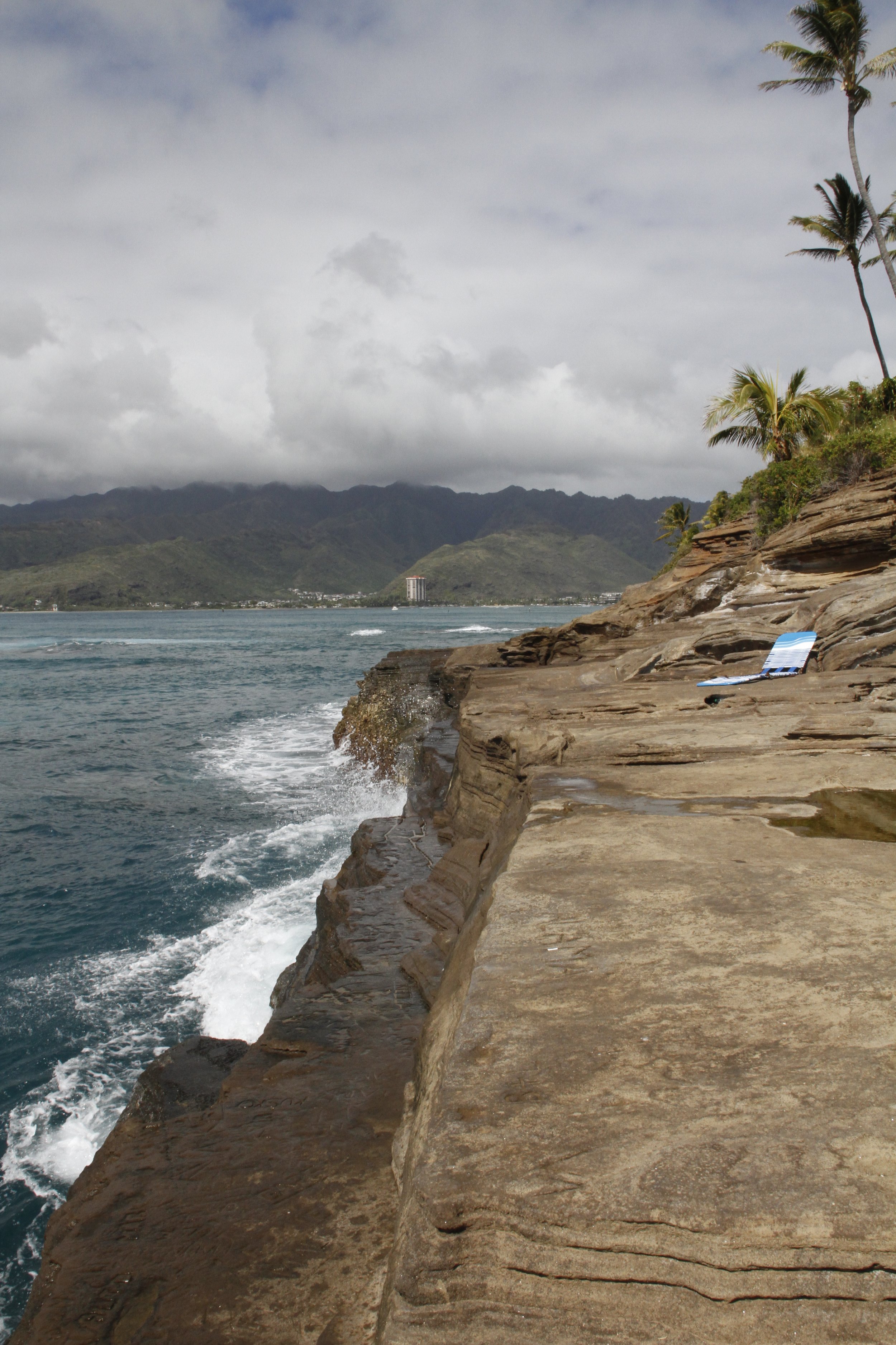 Rocky seaside cliff with ocean waves and distant mountains, palm trees, and a blue beach chair on the rocks. Hawaii film, video, photo production service