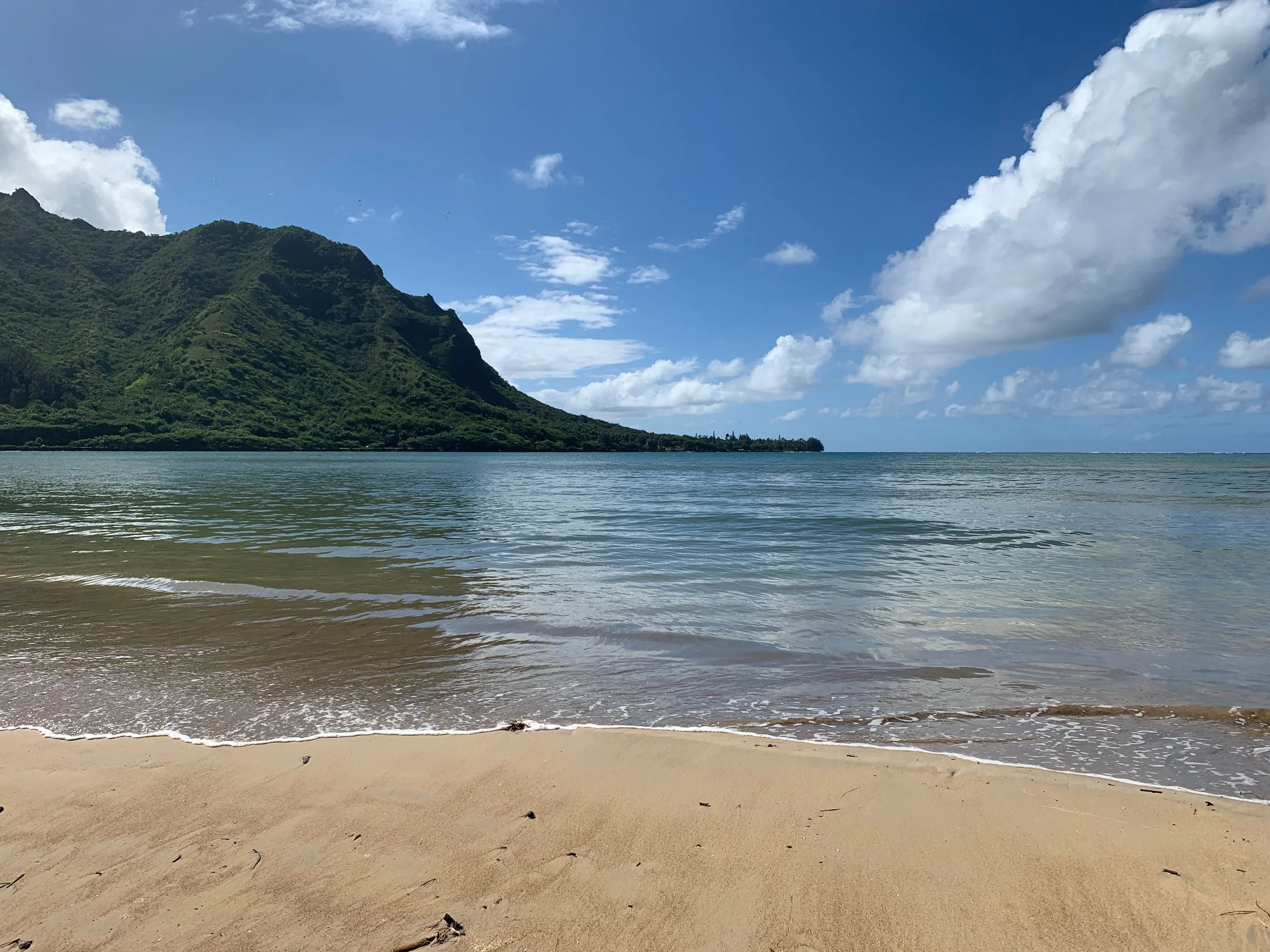 Sandy beach with calm ocean waves, a mountainous green landscape in the background, and a bright blue sky with fluffy white clouds. Hawaii film, video, photo production service