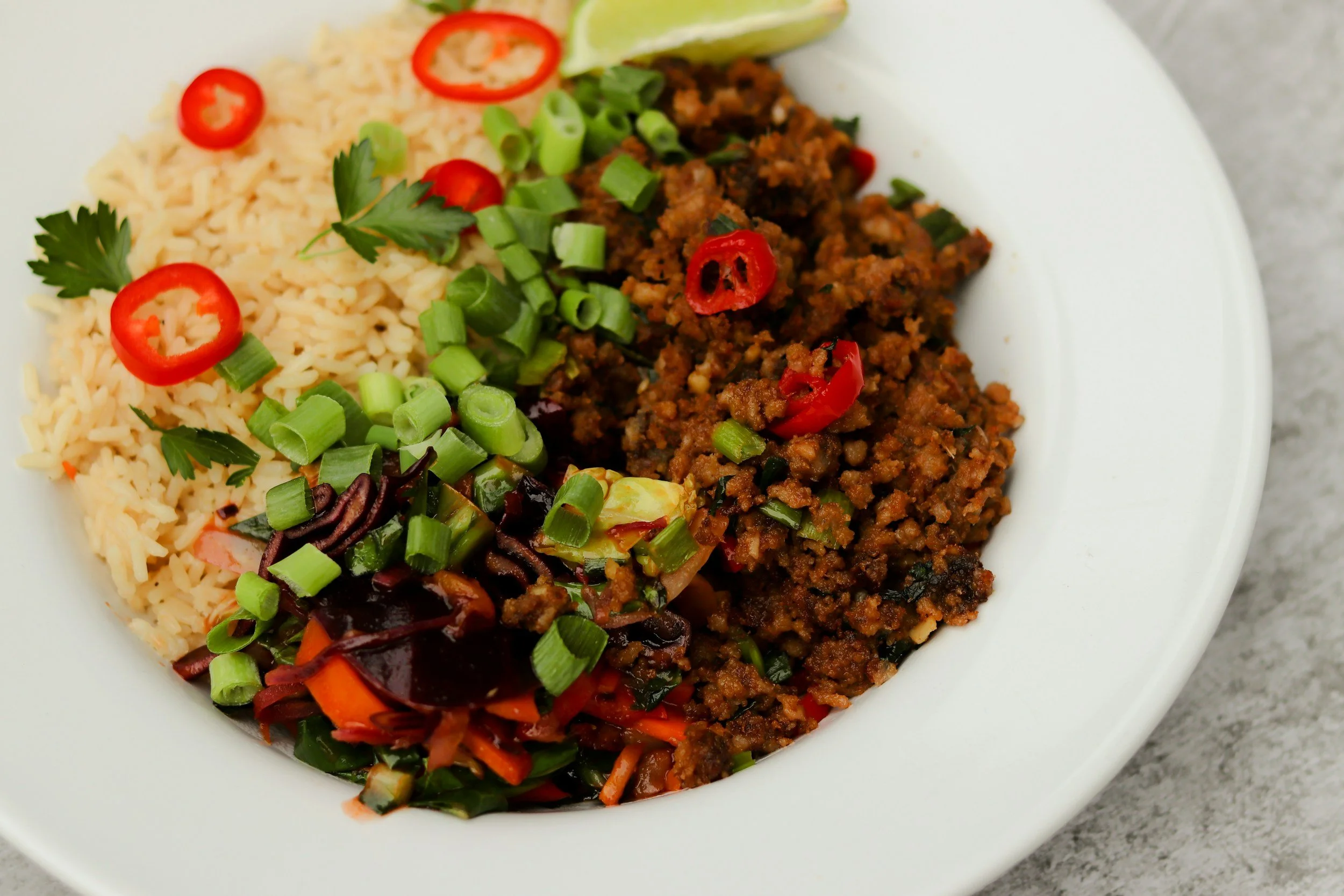Plate of seasoned ground beef, rice, chopped green onions, sliced red chili peppers, cilantro, and a lime wedge.