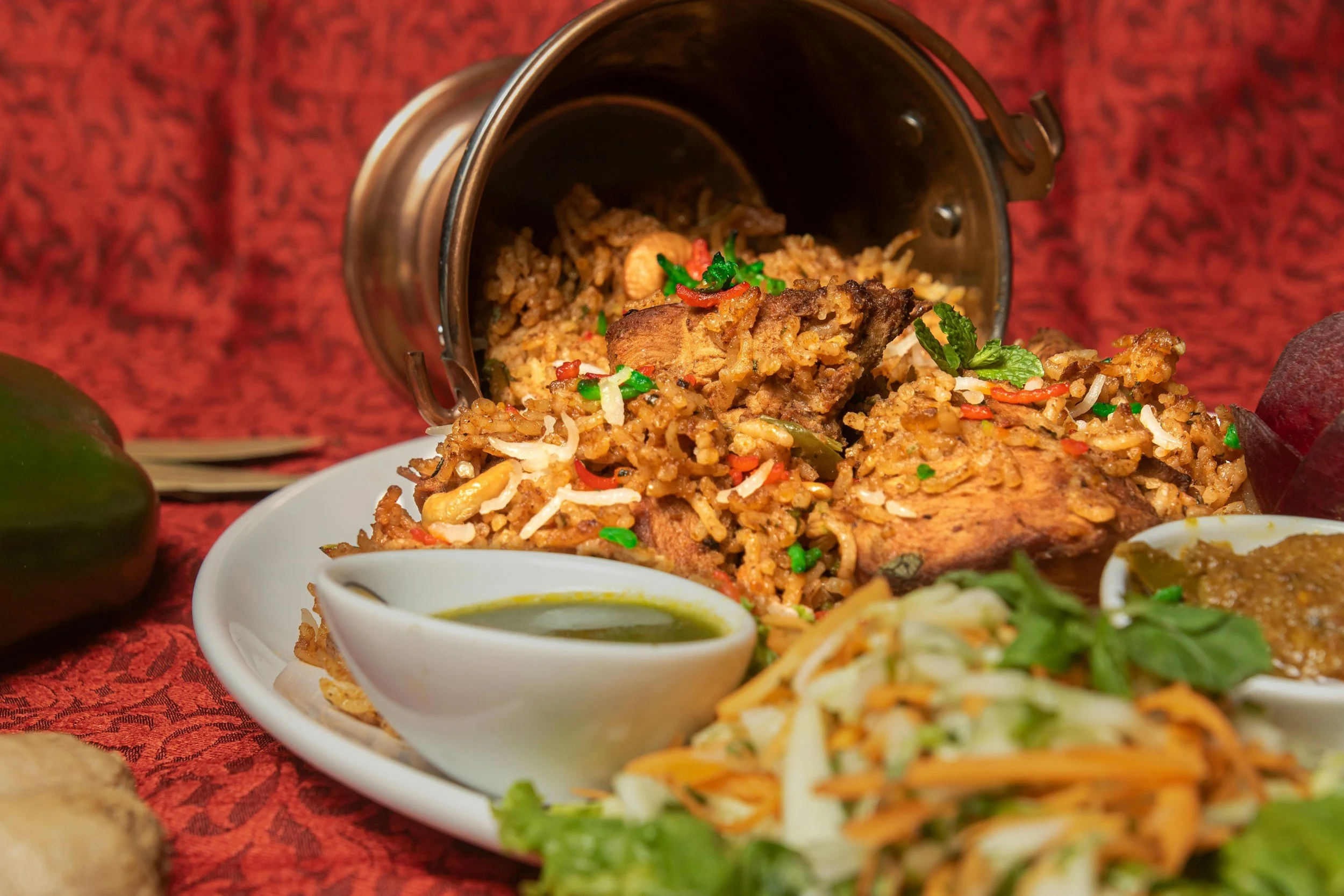 A plate of traditional Indian chicken biryani served with green chutney, salad, and curry, with a red fabric background.